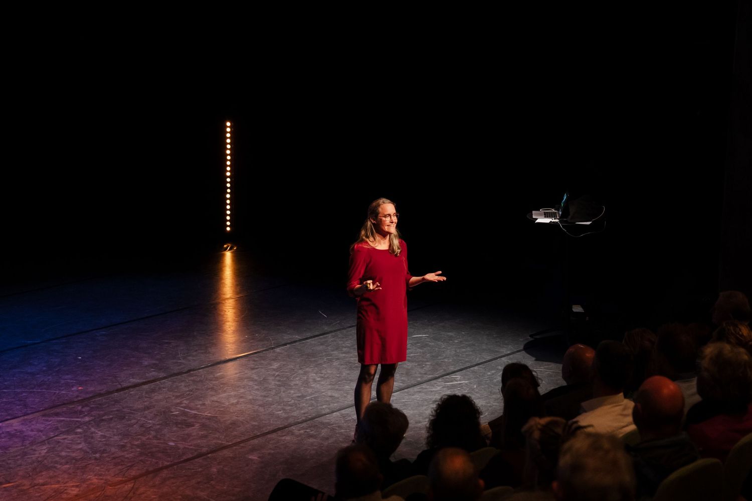 Woman in red dress presenting on stage to seated audience in theater.