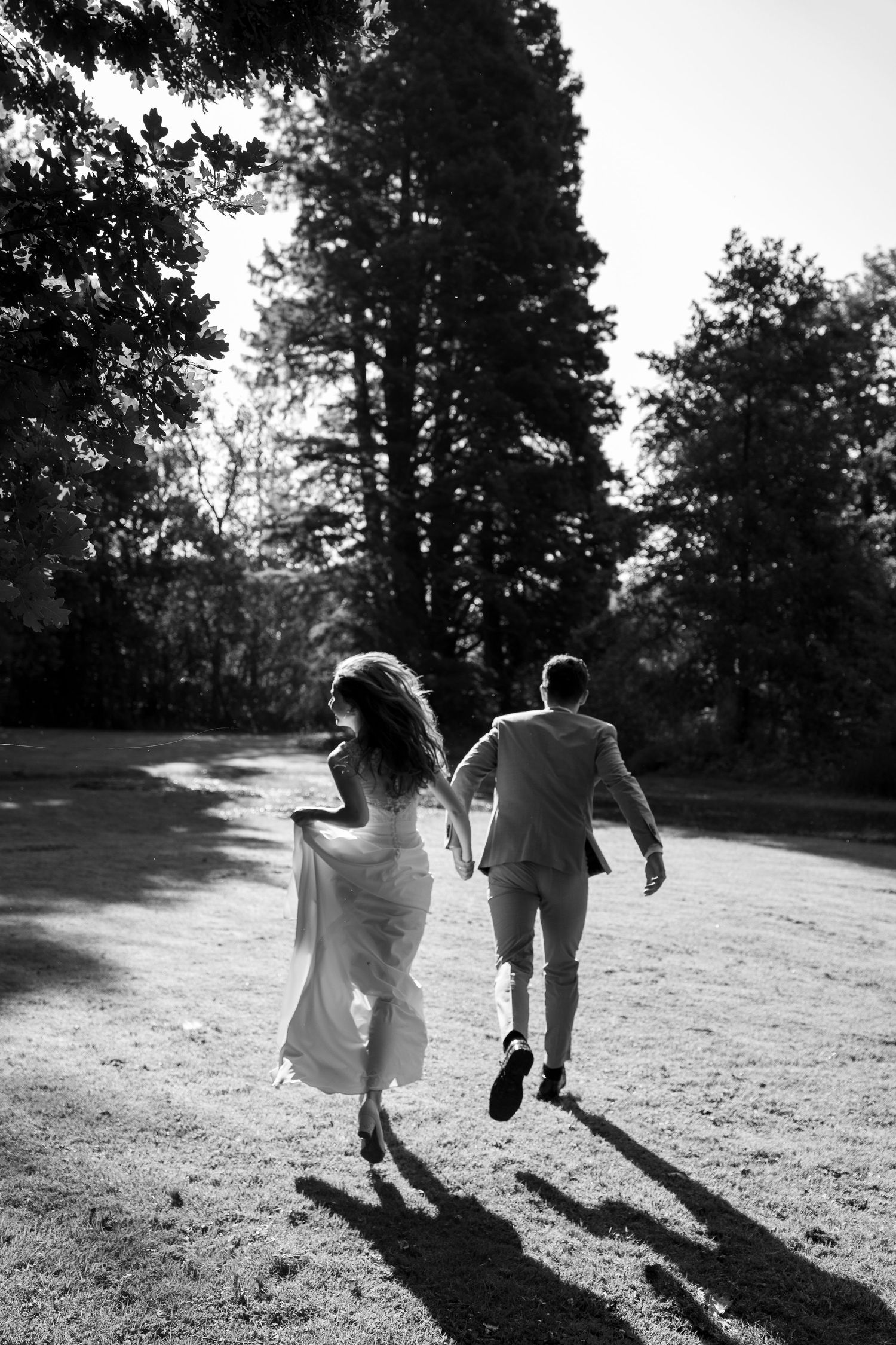 Black and white photo of bride and groom running hand in hand through a sunlit park on their wedding day.