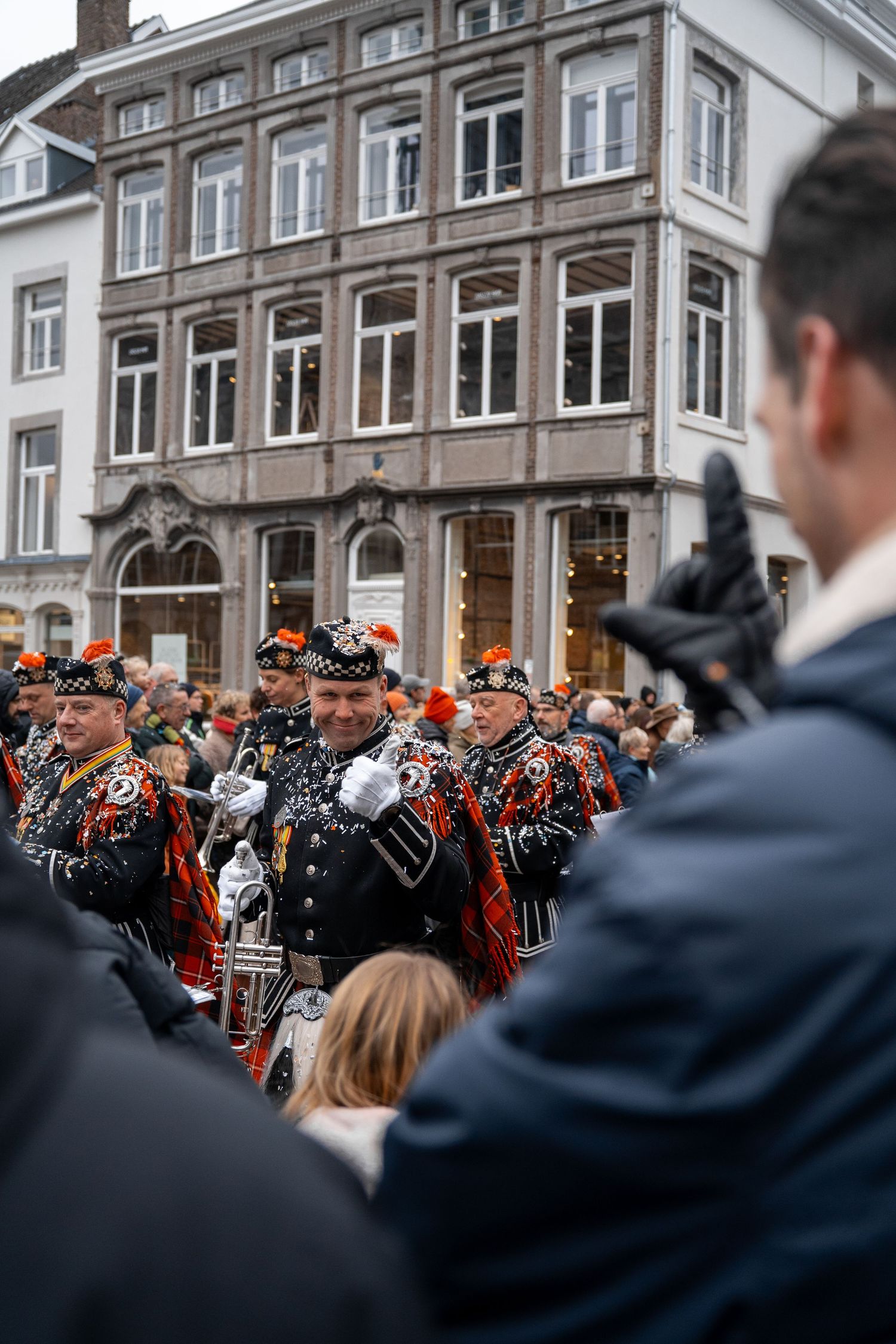 Marching band members in black ornate uniforms with red feathered hats performing in a winter street parade.