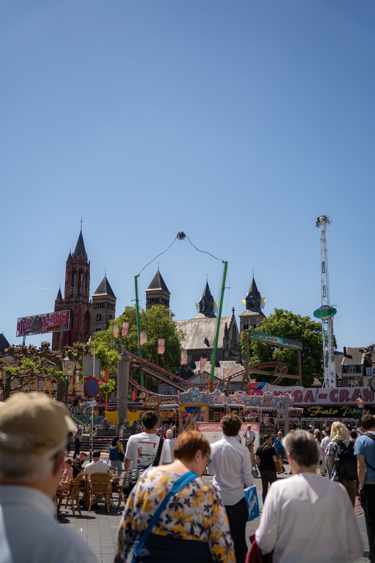 Crowds of people enjoying a sunny summer fair with colorful rides and a historic church visible in the background.