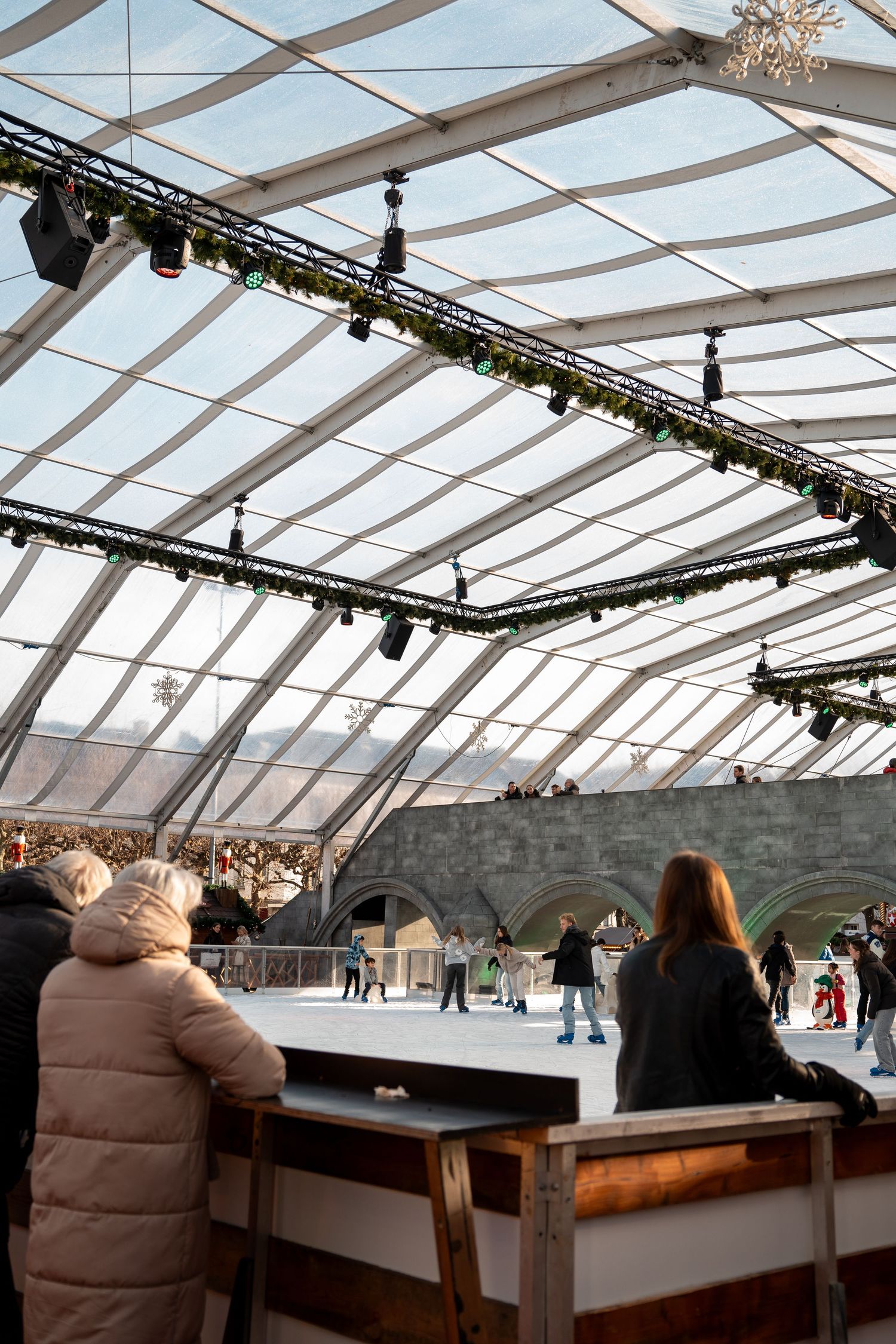 Indoor ice skating rink under a glass roof with spectators watching skaters during winter holidays.