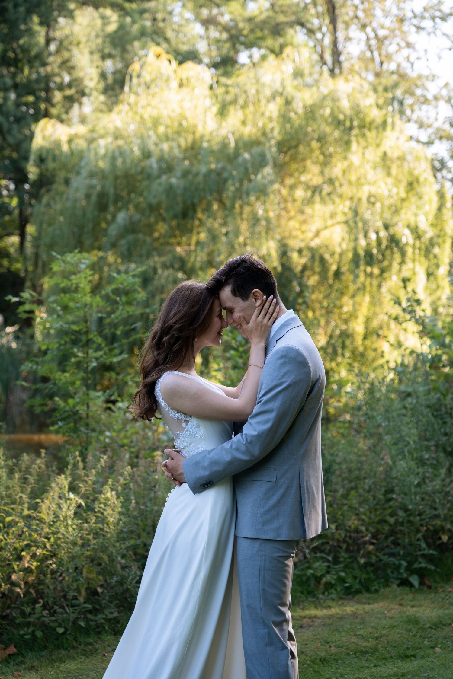Romantic wedding couple embracing forehead to forehead in a lush green garden with soft golden light.