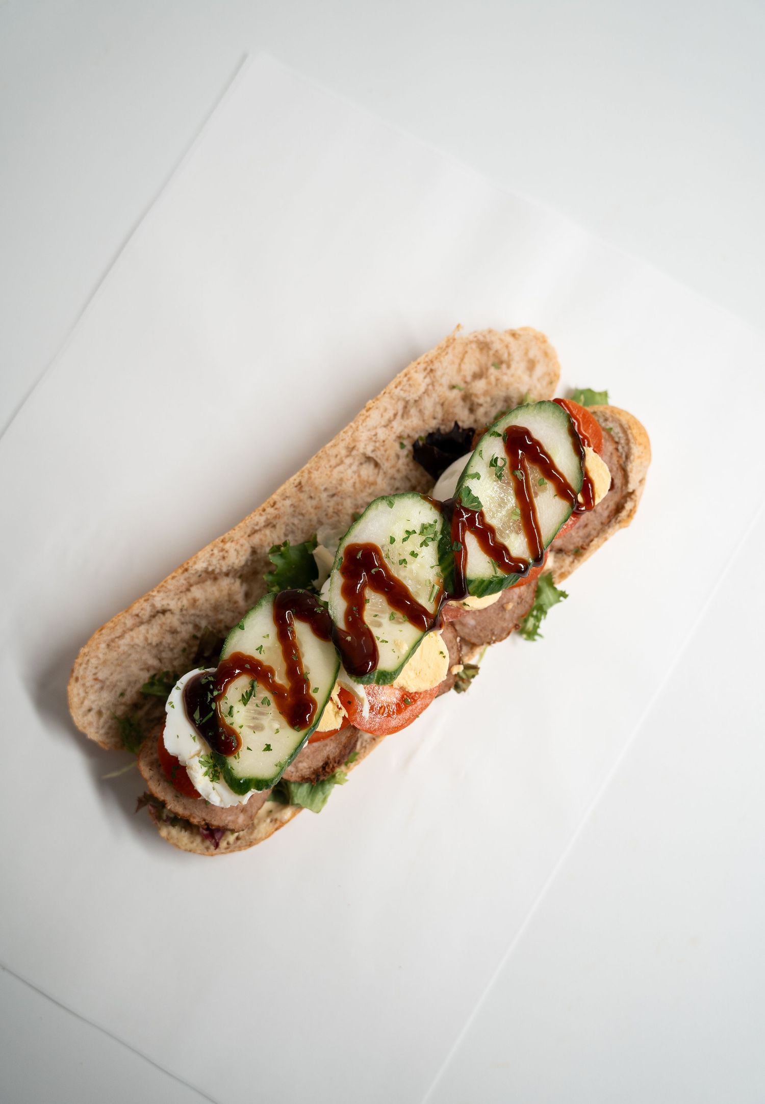 A gourmet open-faced sandwich on a hoagie roll topped with vegetables, egg, and drizzled BBQ sauce on white paper.