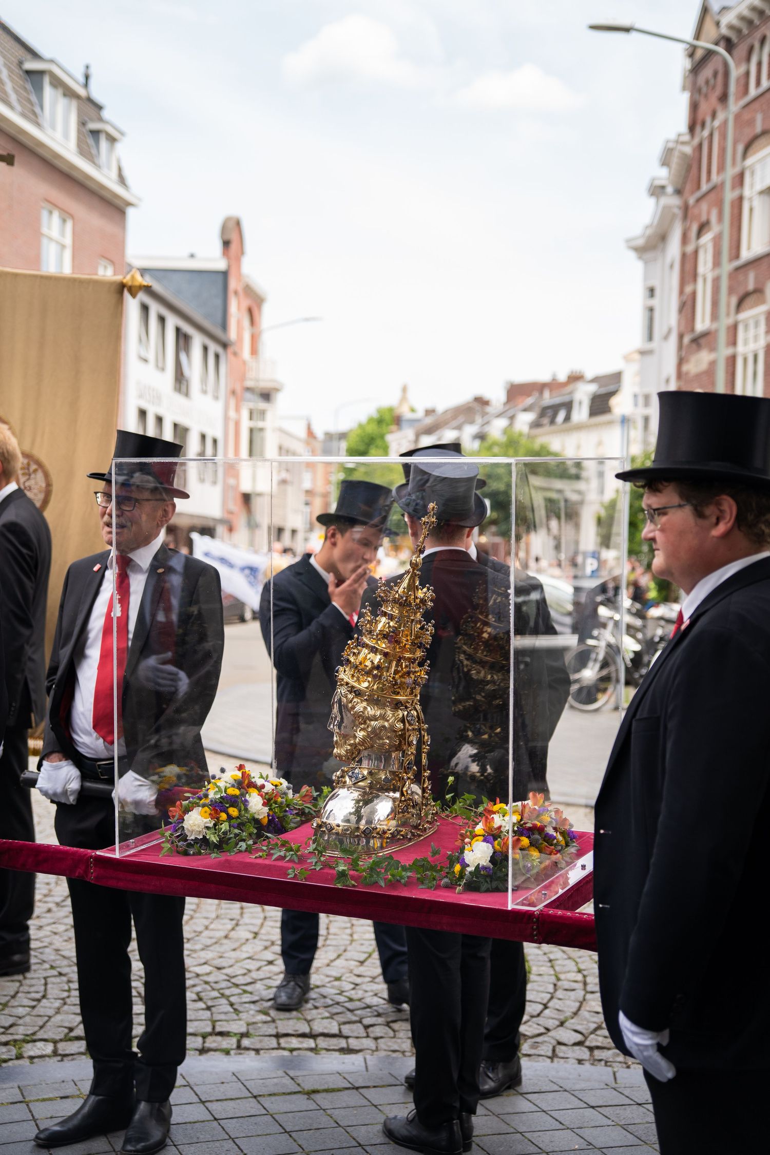 Formally dressed men in top hats carry golden religious relic in glass display case.