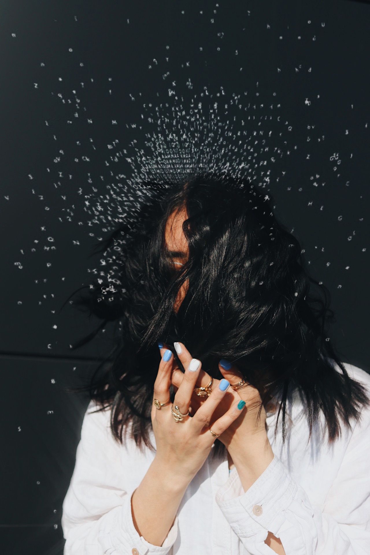 Woman with dark hair covering face shaking head with water droplets flying in sunlight.
