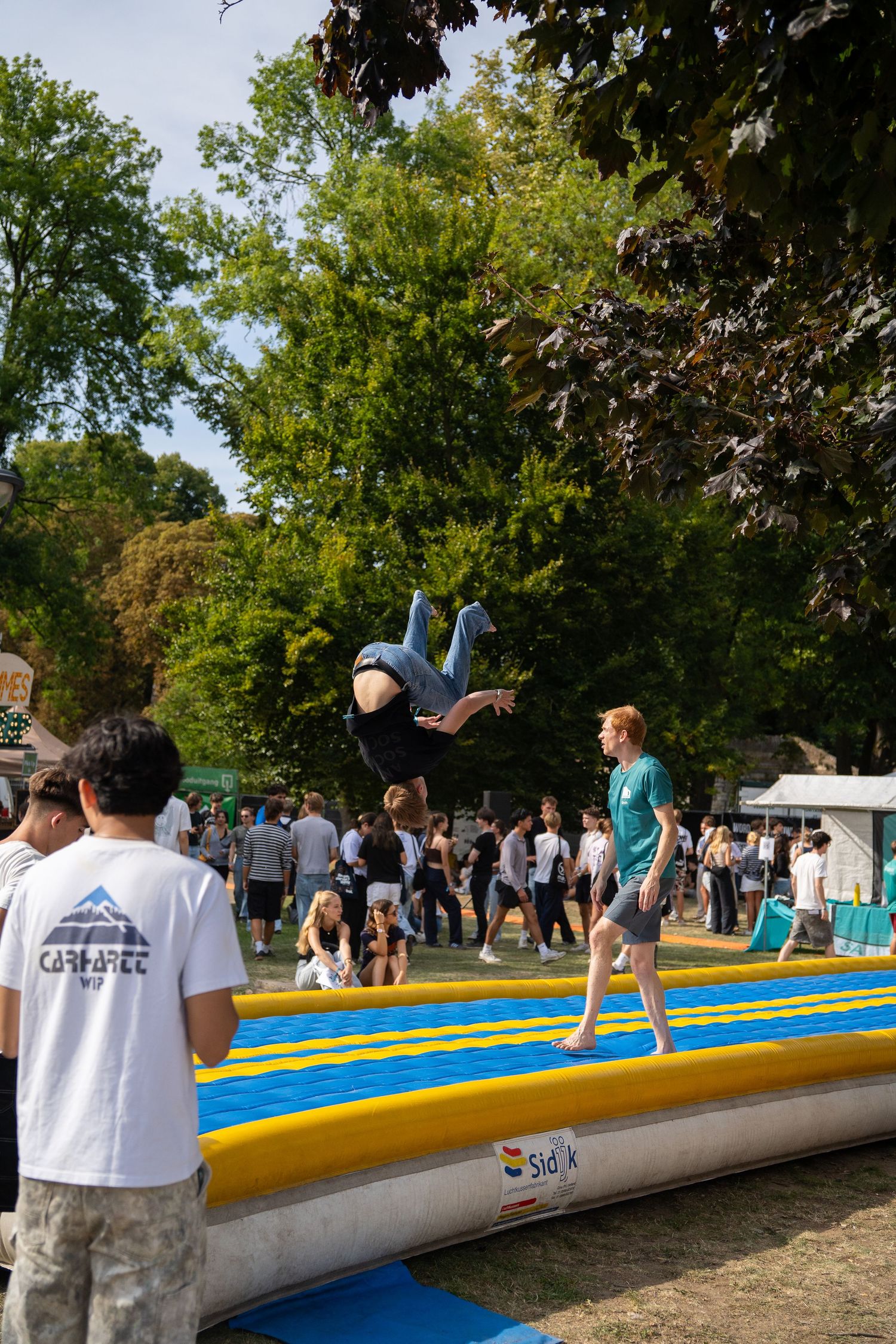 Person performing a backflip on an inflatable air track at a lively outdoor summer festival.