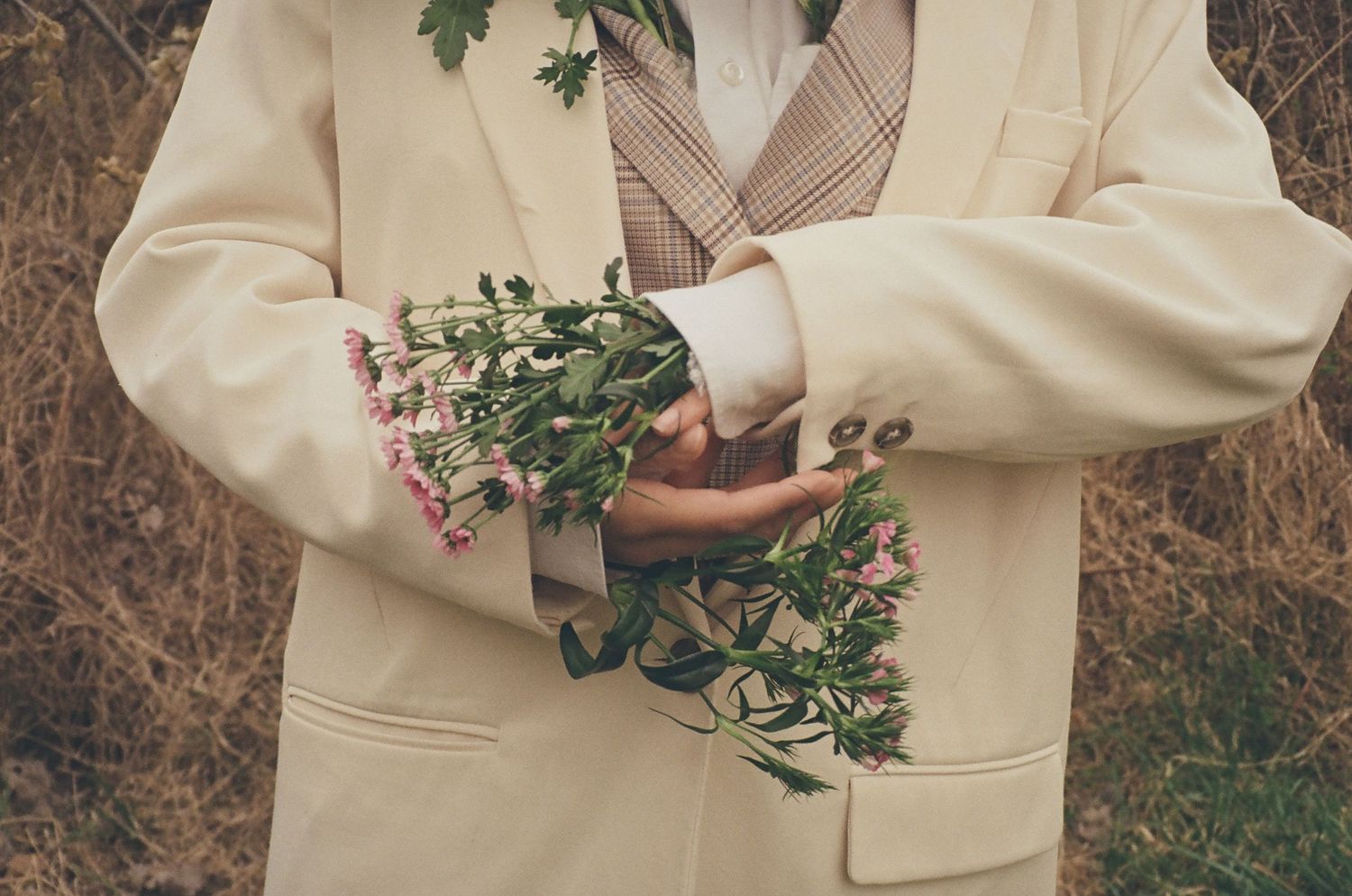 Person in cream blazer holding wildflowers with crossed arms against a dry grass background.