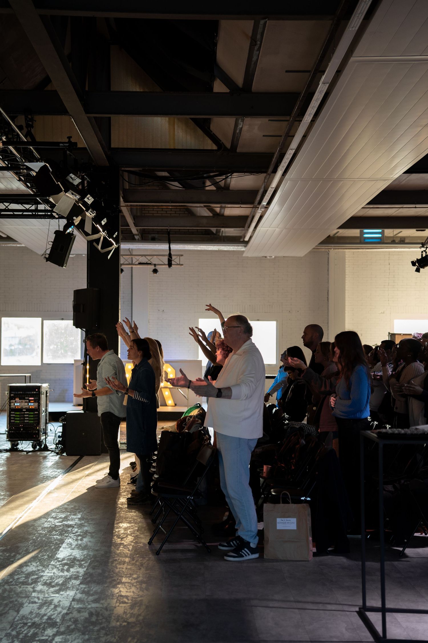 Congregation members raising hands in worship during a sunlit church service in an industrial-style venue.