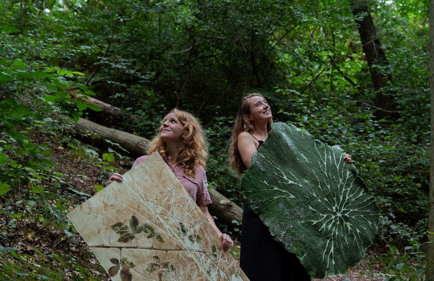Two women carrying large botanical leaf artworks through a lush green forest setting.