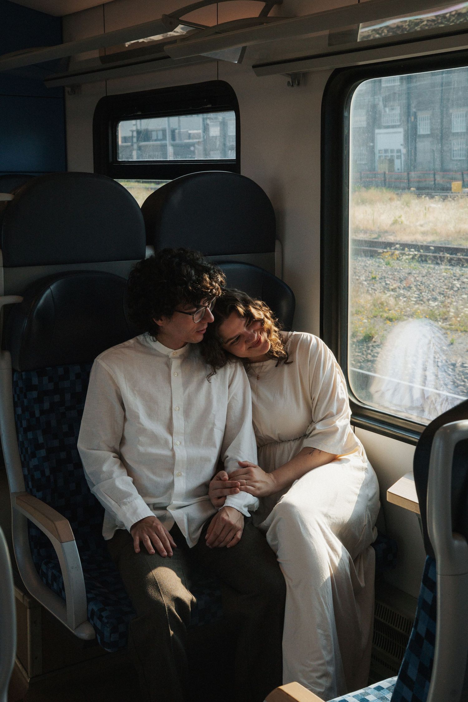 A couple in white clothing sits closely together on a train, the woman resting her head on the man's shoulder.