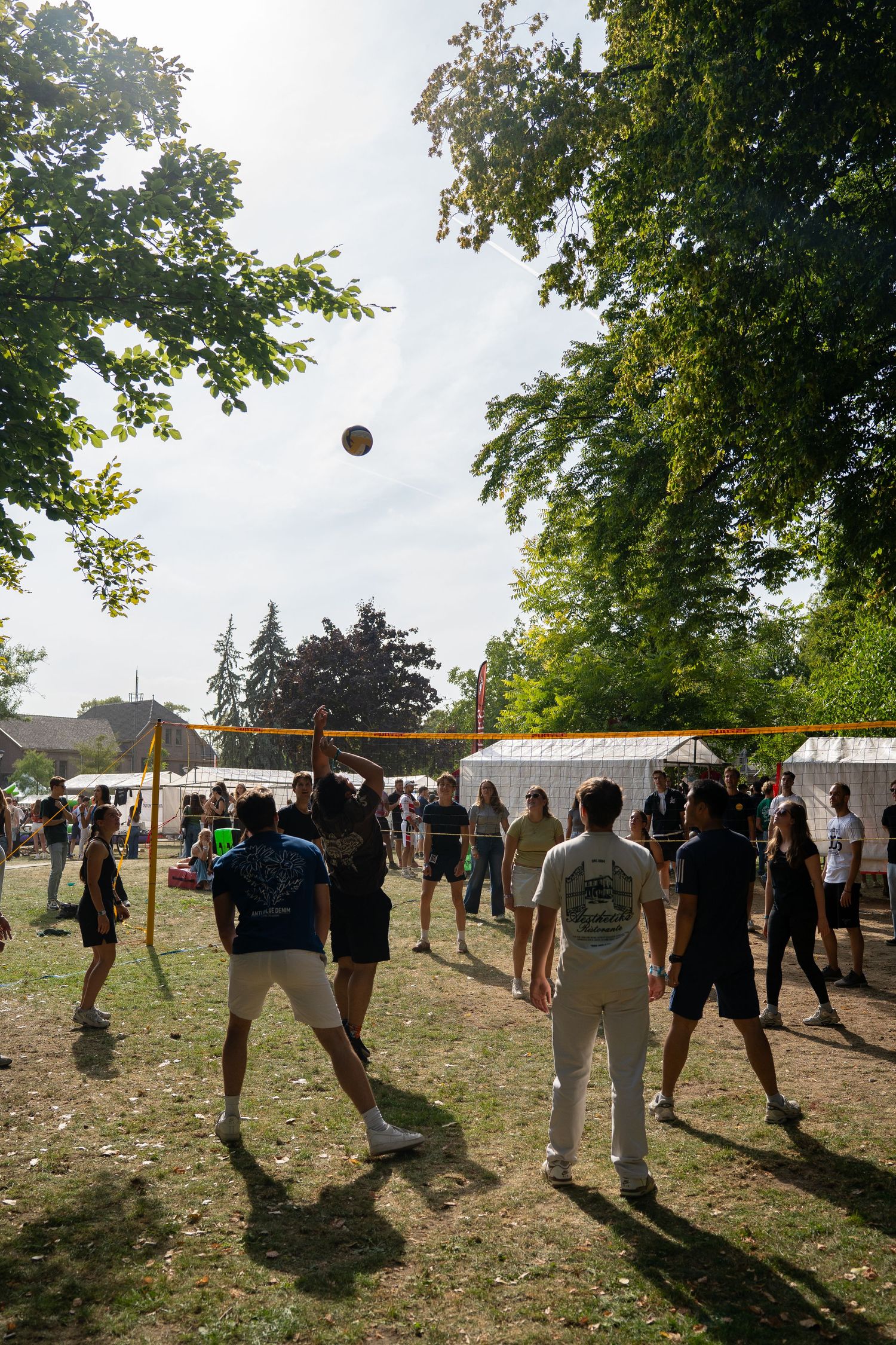 Group of young people playing volleyball outdoors at a sunny summer festival with white tents in the background.