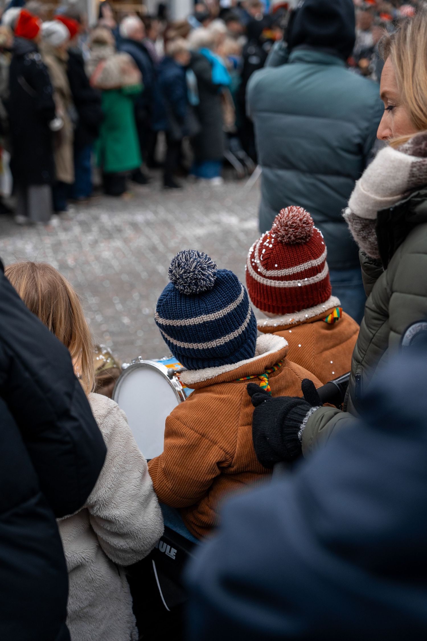 Two young children in colorful winter pom-pom hats watching a crowded outdoor winter parade with their mother.