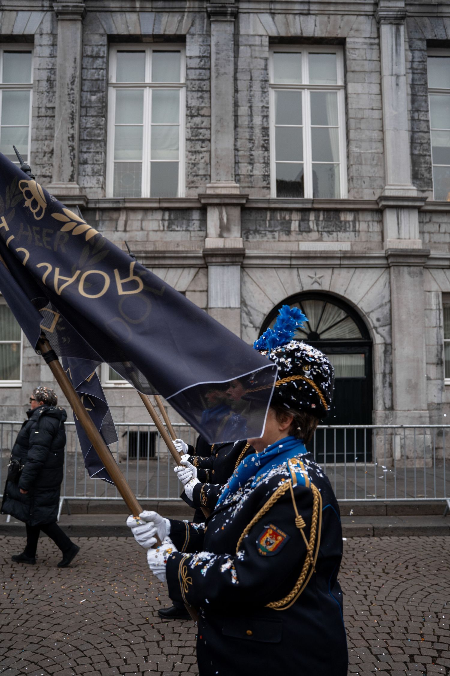 Parade performer in uniform carrying a dark ceremonial flag dusted with snow outside a historic stone building.