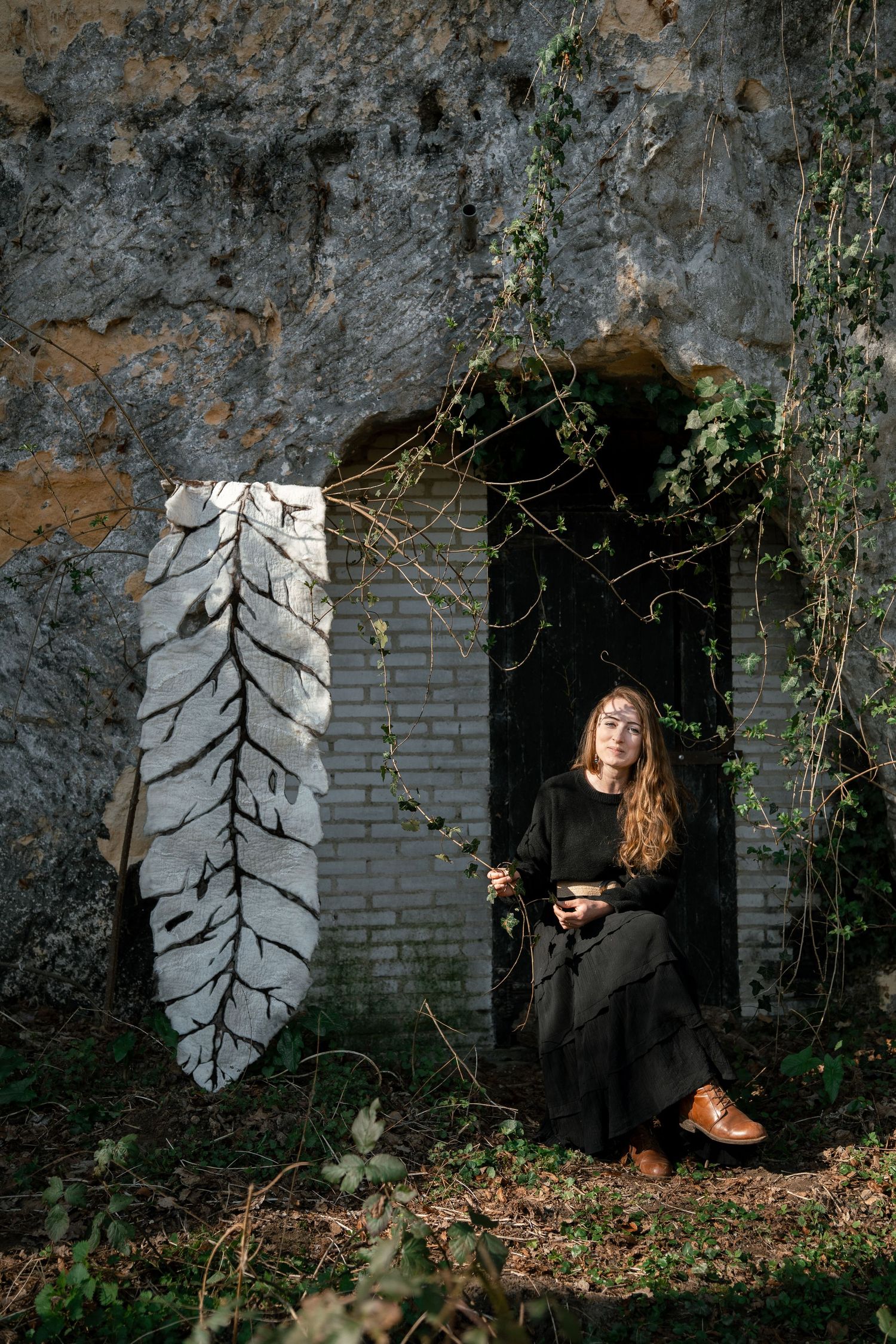 Woman in black dress sits in a doorway beside a large leaf sculpture on a moss-covered stone wall.