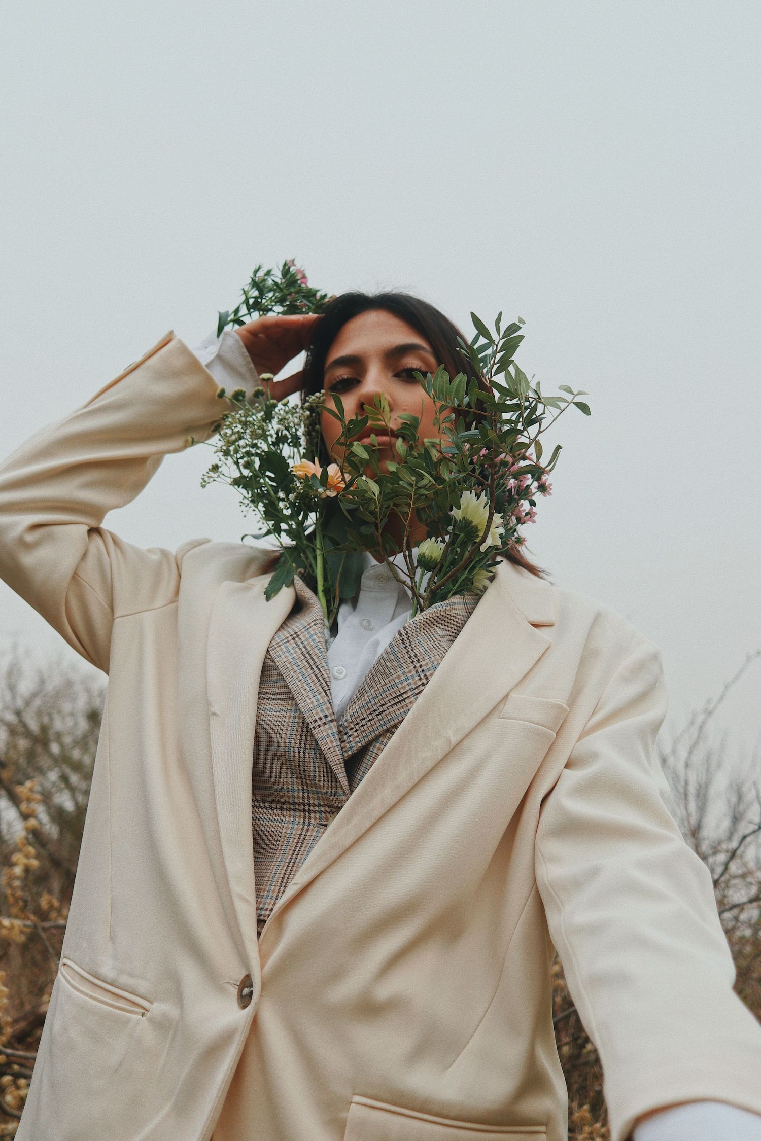 Person in beige blazer and plaid suit holds wildflowers covering their face against a muted autumn outdoor backdrop.