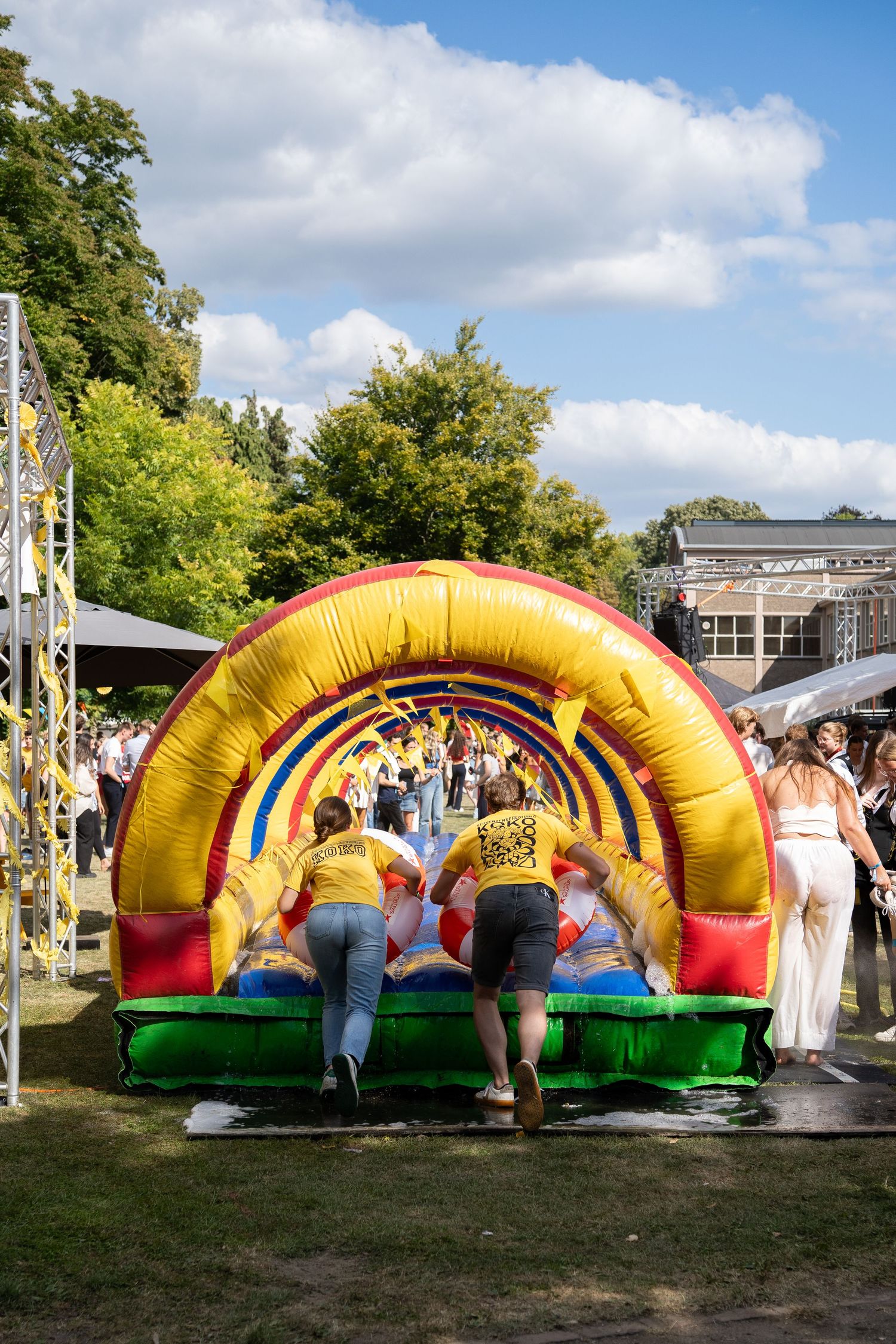 Two people climbing into a colorful inflatable bounce house slide at a sunny outdoor festival.