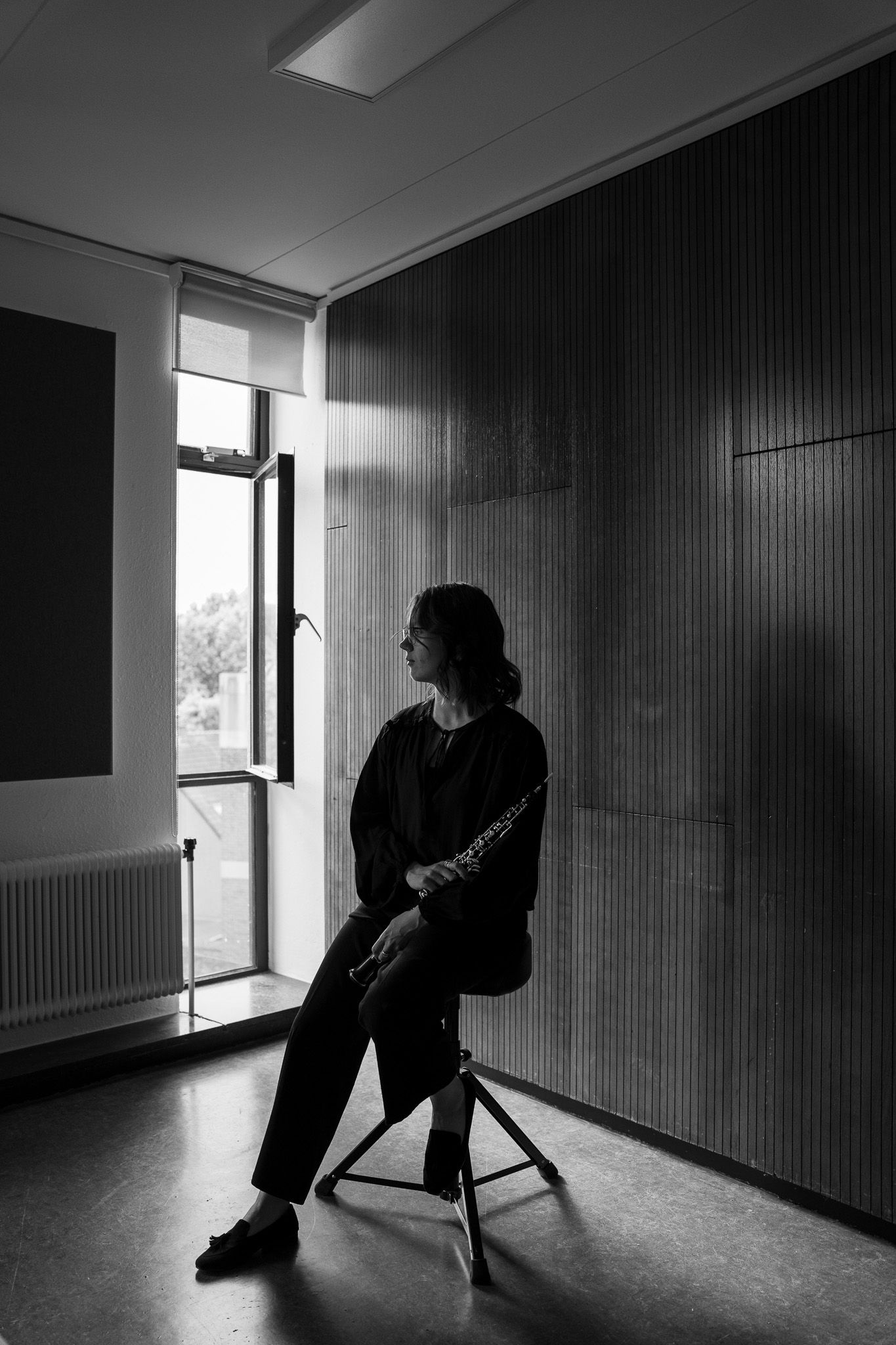 Black and white silhouette of a female oboist seated on a stool near a window holding her instrument.