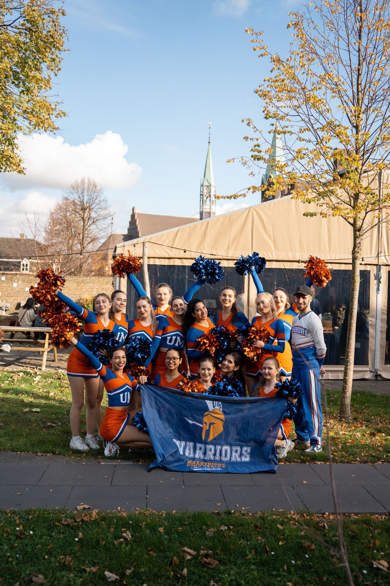 Maastricht University Warriors cheerleading squad poses outdoors with blue and orange pom-poms and banner.