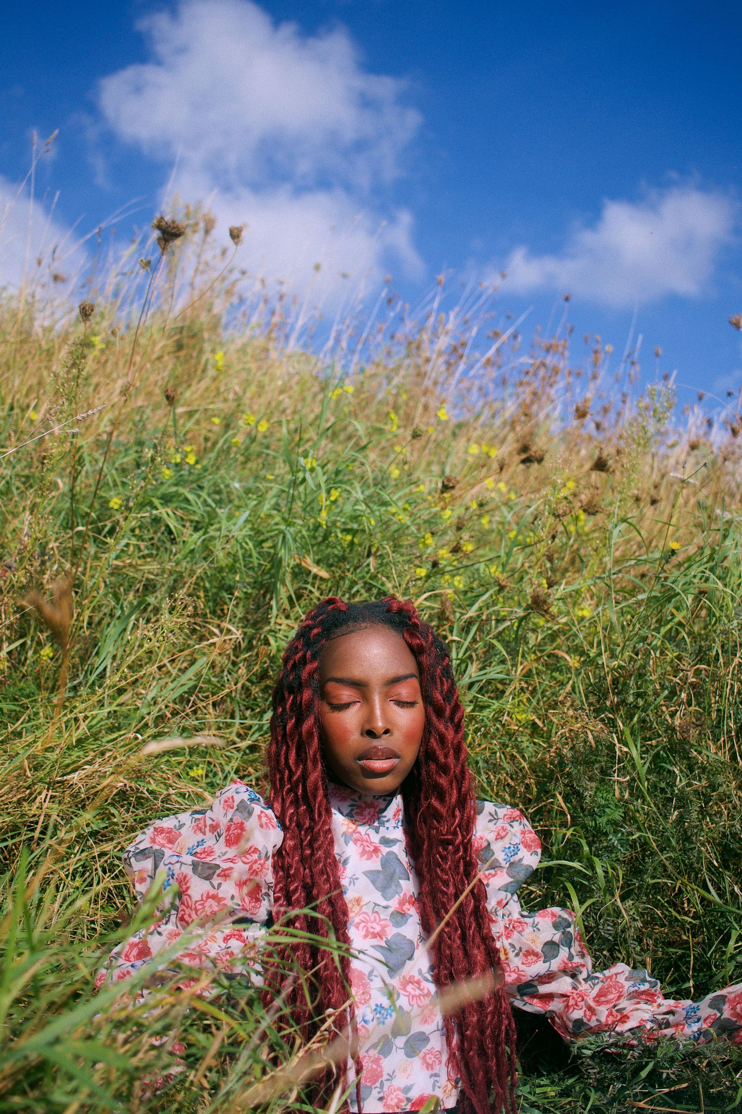 Young Black woman with red braids in floral outfit sits peacefully in tall grass under blue sky.