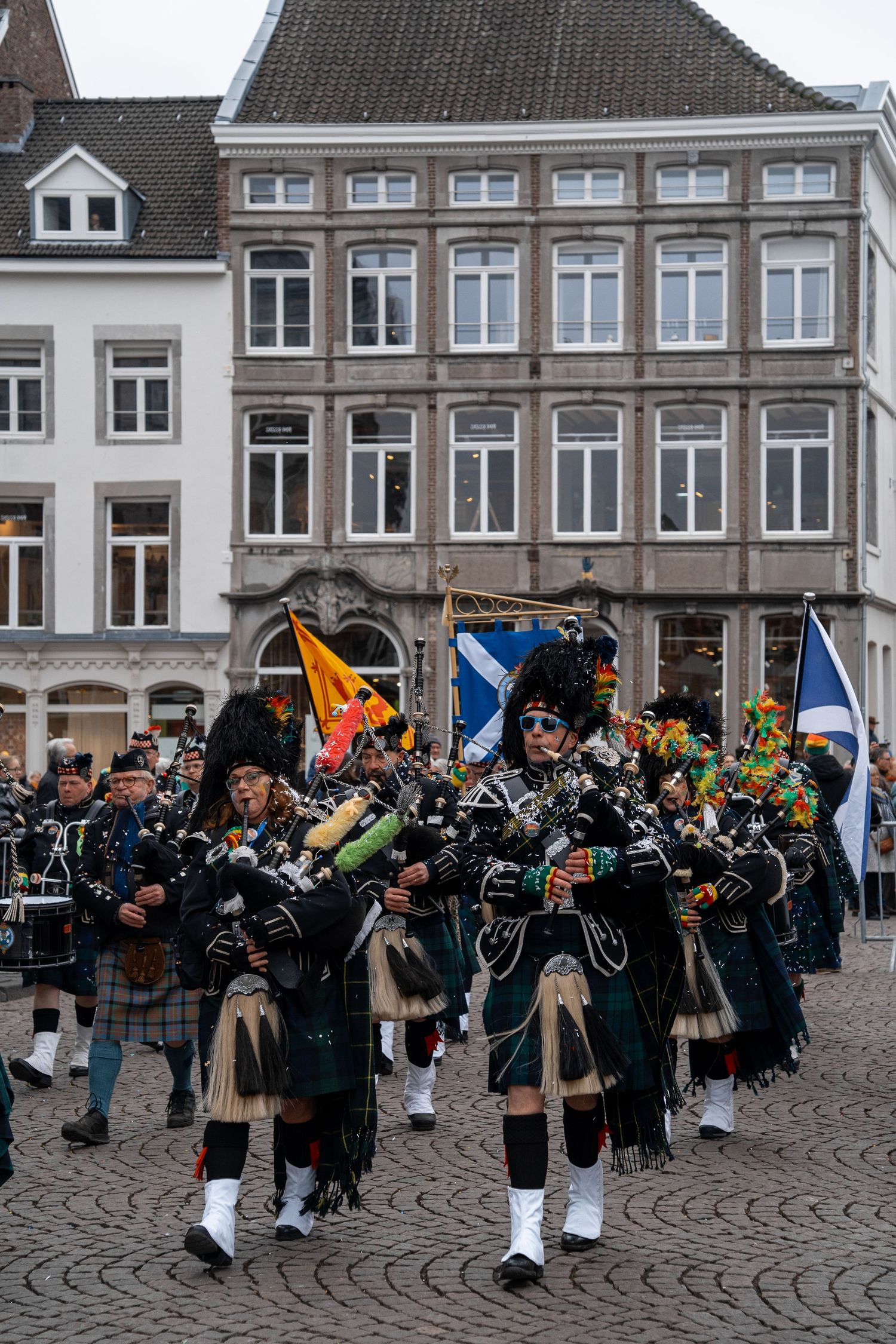 Scottish bagpipe band in traditional kilts marching through a European cobblestone square during a festive parade.