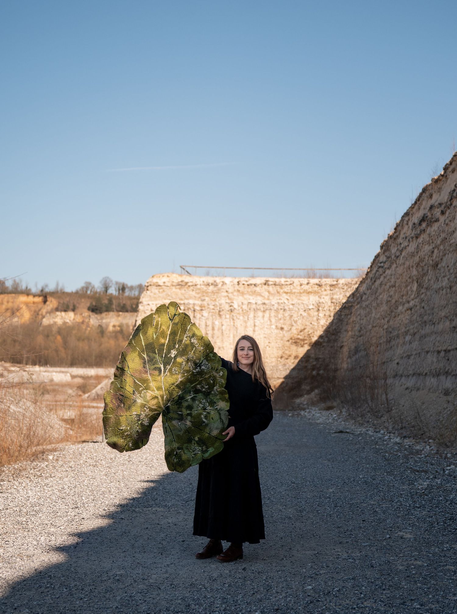 Woman in black dress holding large decorative leaf fan in rocky quarry landscape.