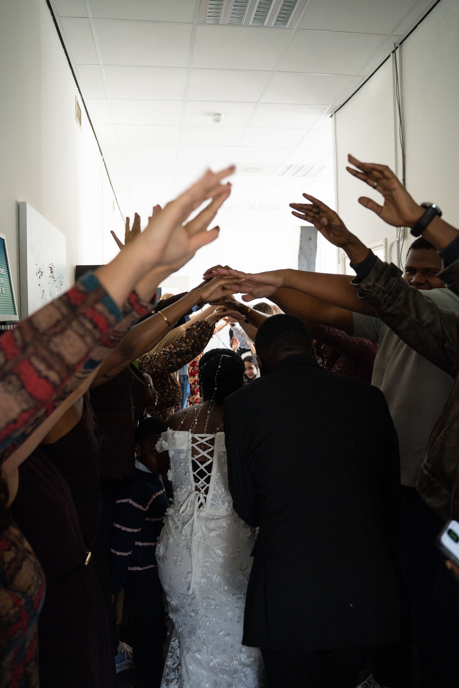 Wedding guests raise hands in blessing over bride and groom in hallway ceremony.
