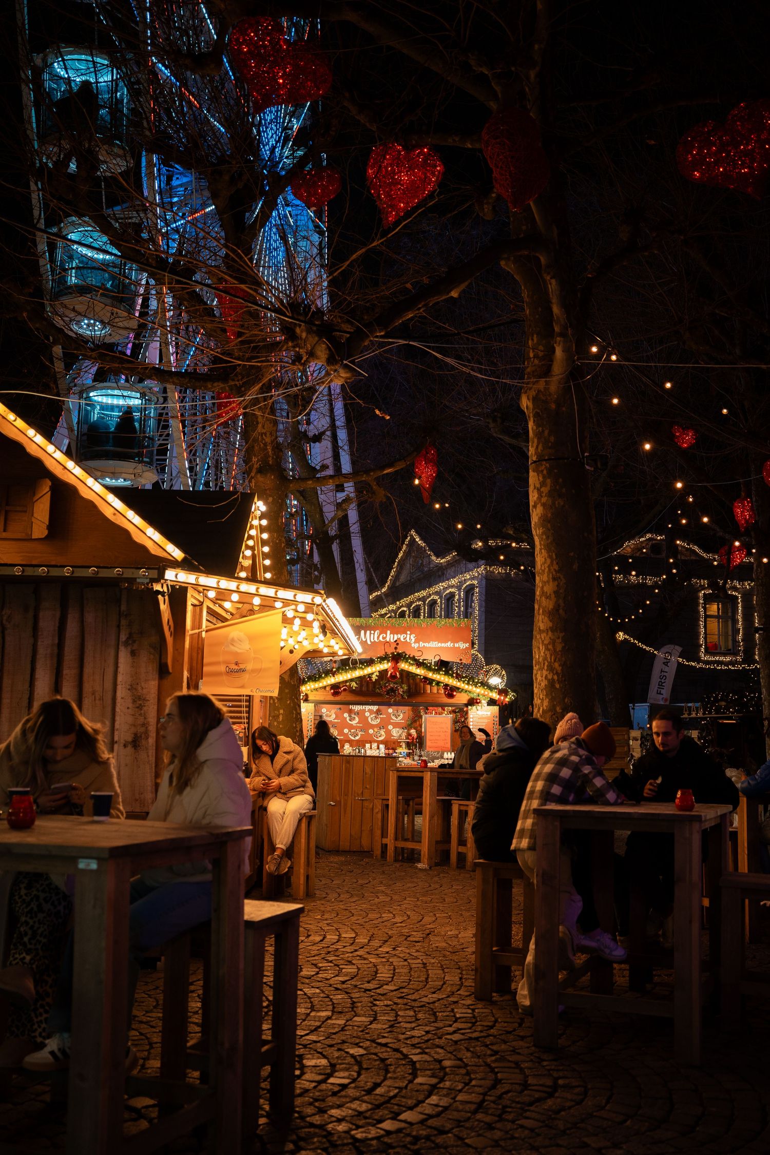 Festive Christmas market at night with glowing stalls, red heart decorations, and a Ferris wheel.