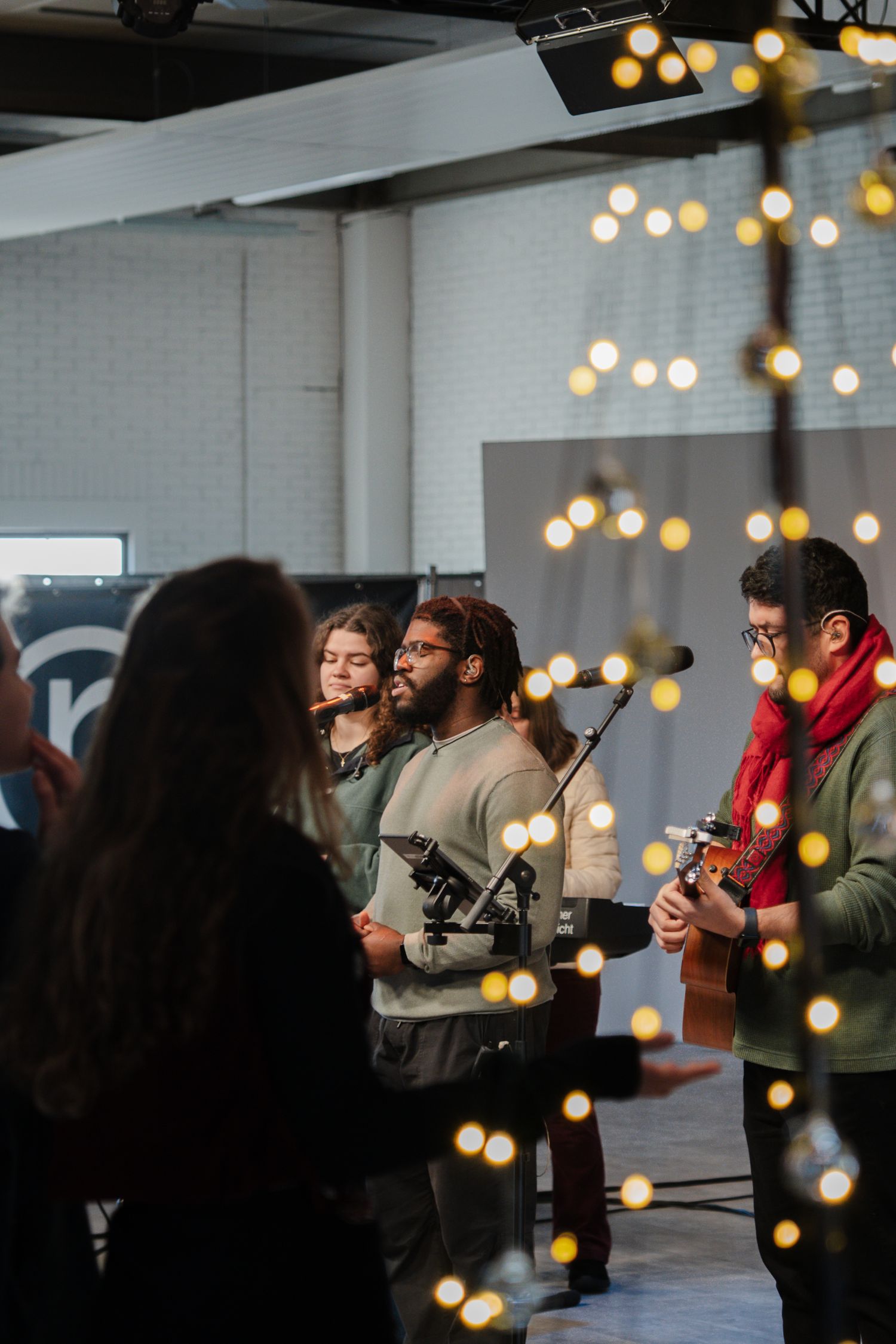 Worship band performing with guitars and microphones surrounded by warm string lights in a modern church.