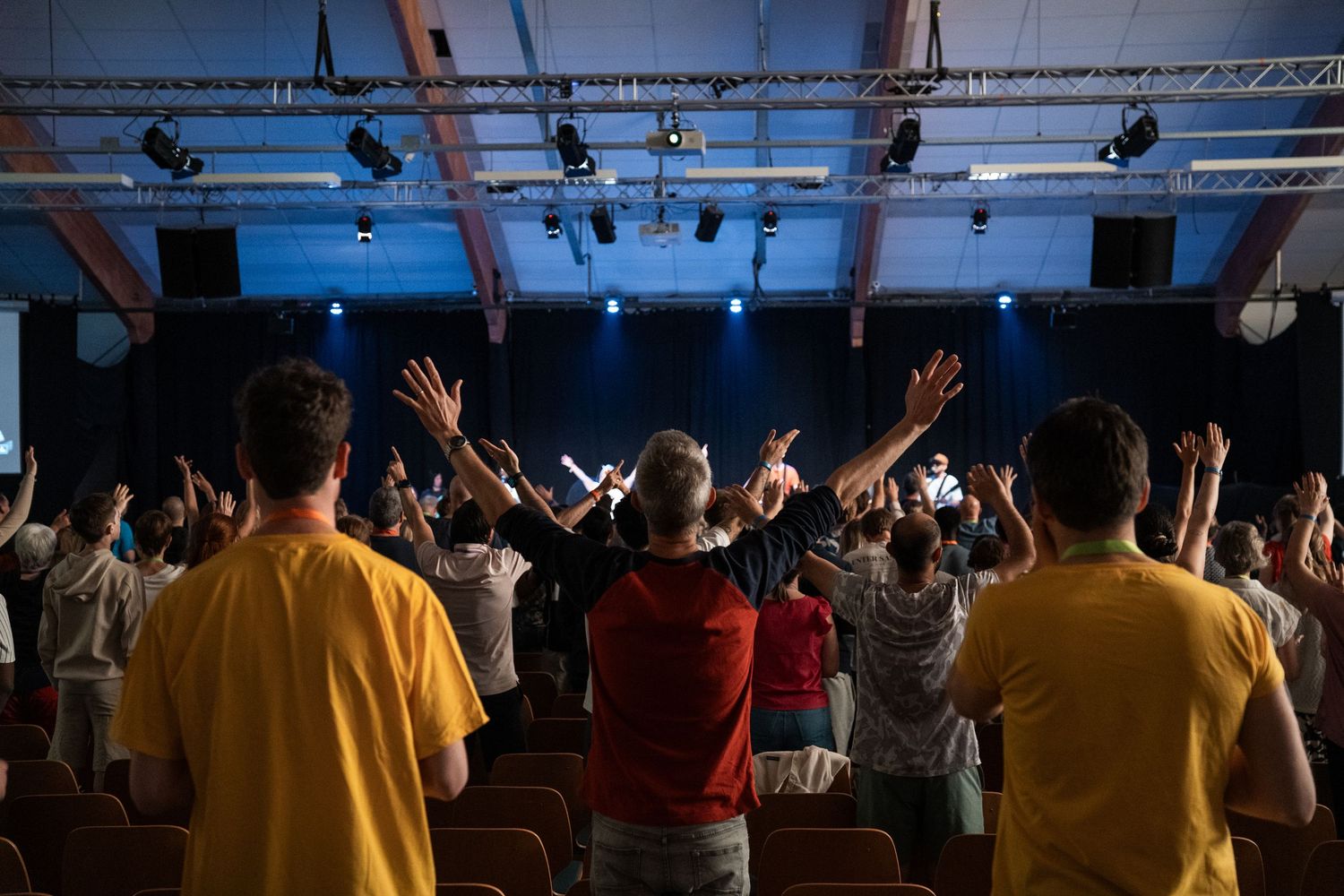 Congregation raising hands in praise toward a blue-lit stage during a worship service.