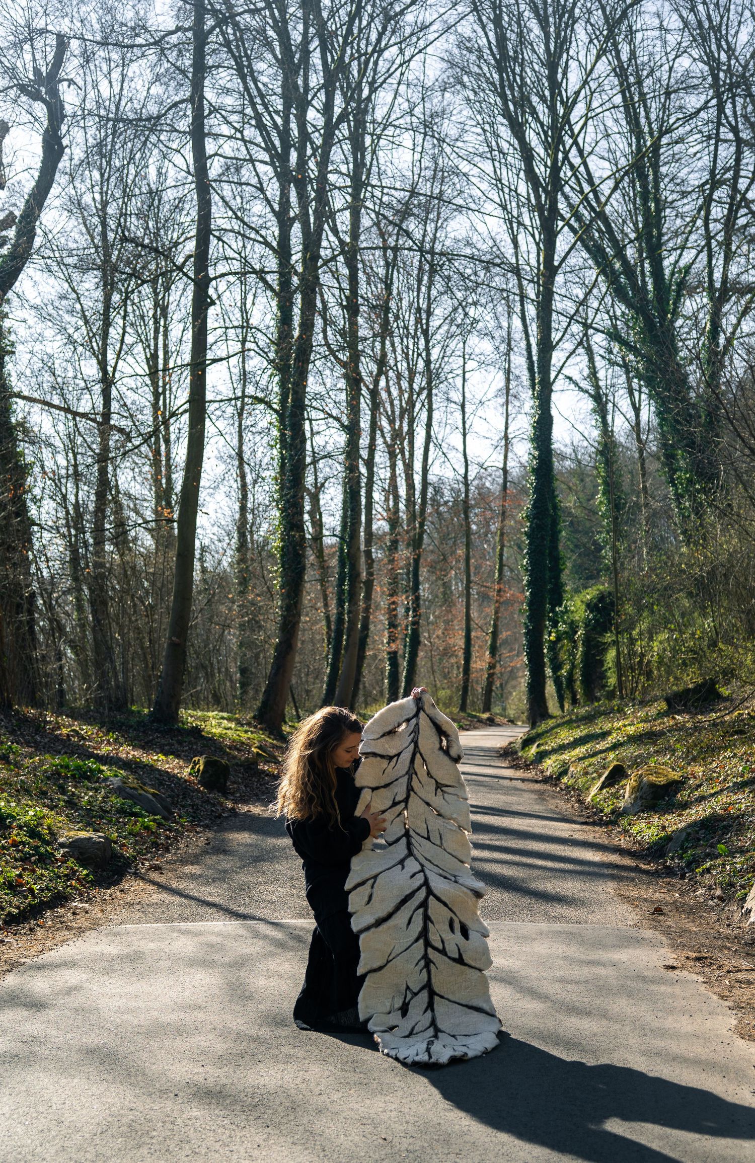 Woman in black dress hugging a large white leaf sculpture on a tree-lined road in winter.