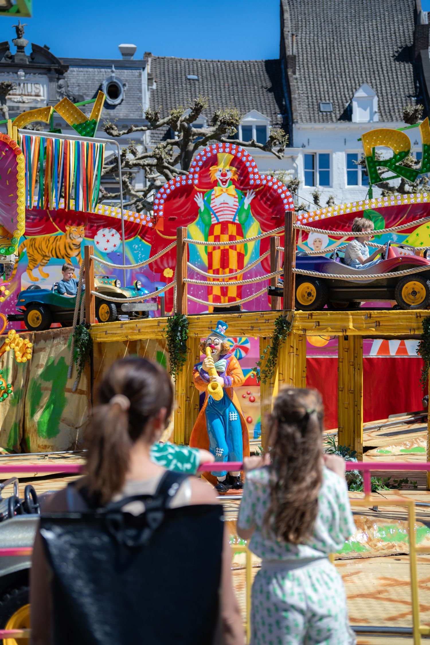 Two girls watch a vibrant circus-themed carousel ride featuring a clown figure at a sunny outdoor fair.
