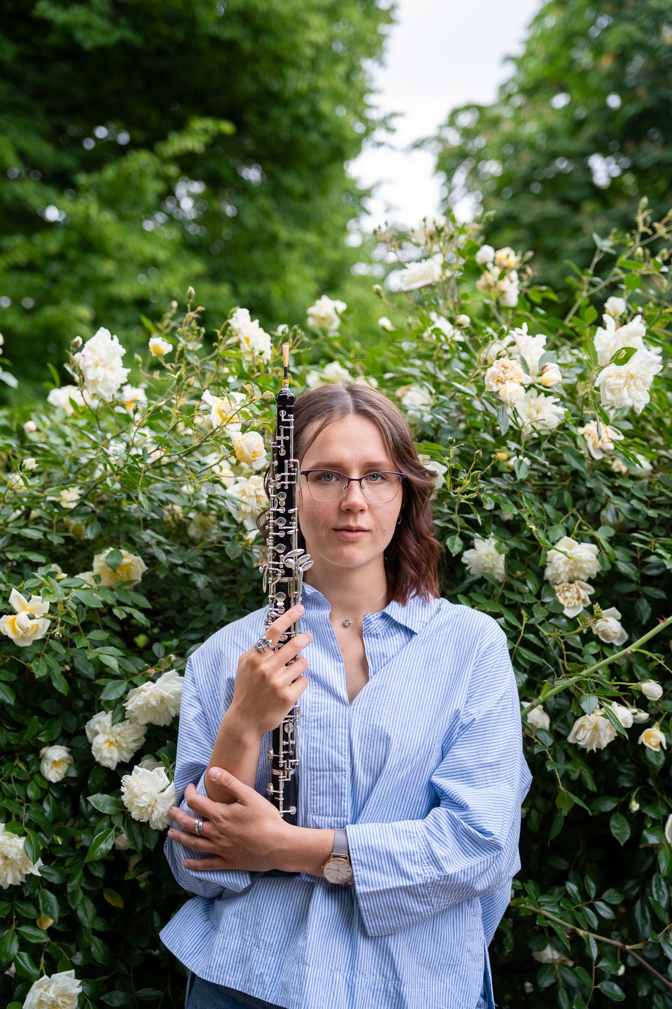 Young woman with glasses holding an oboe instrument in front of blooming white rose bushes.