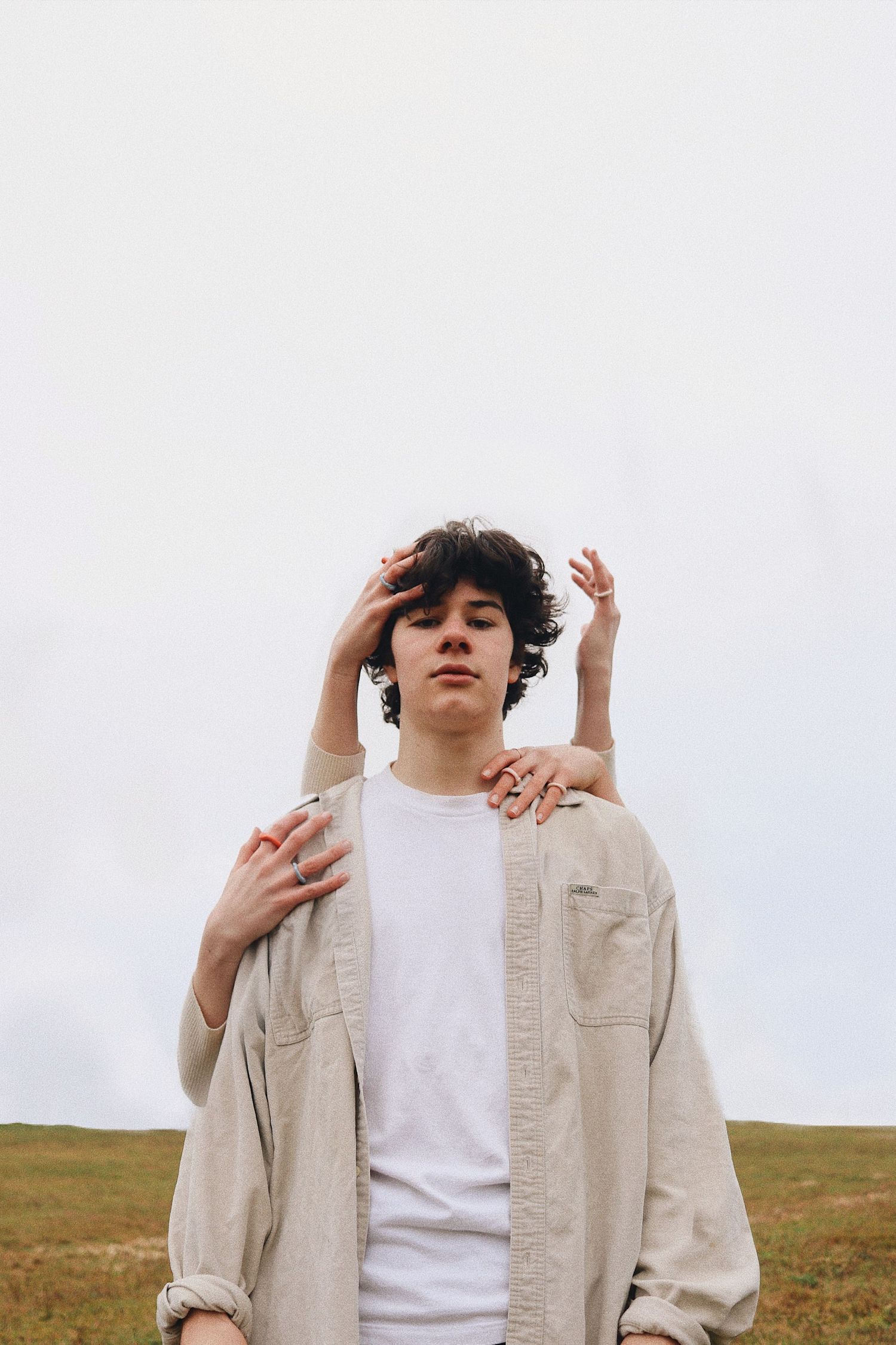 Young man with curly hair in beige shirt standing in a field with hands placed on his shoulders.