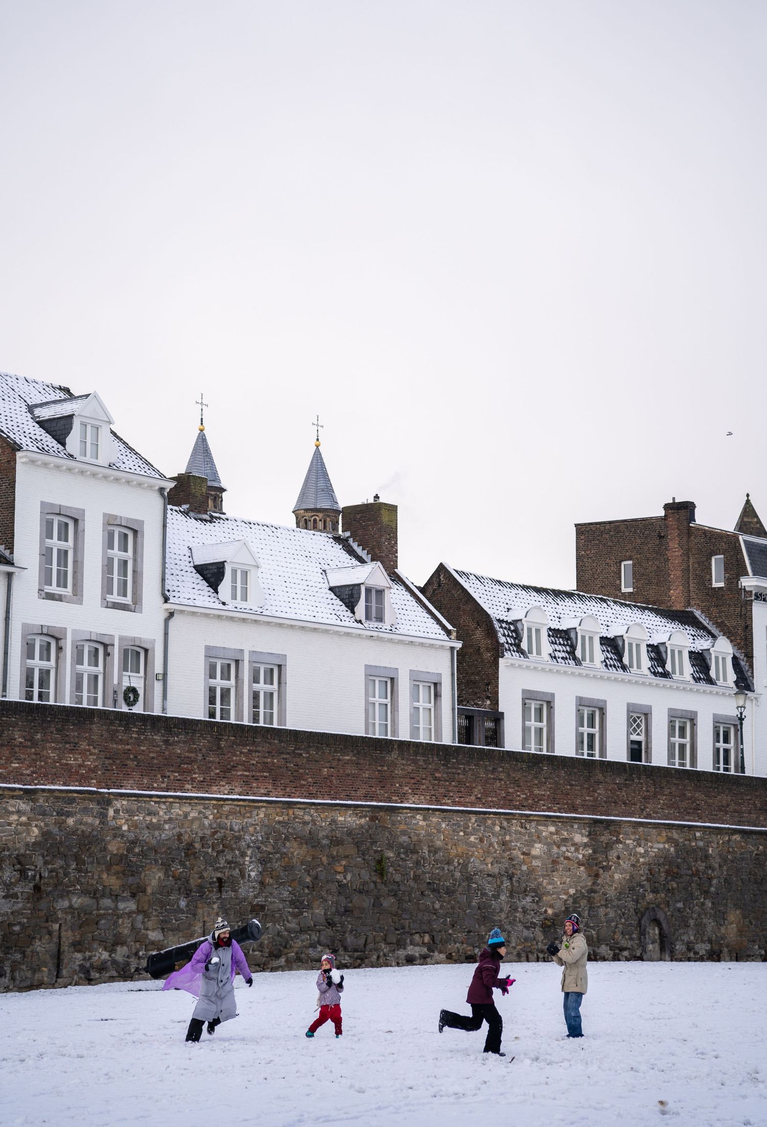 Children playing in the snow near historic white buildings and an old brick city wall in Maastricht.