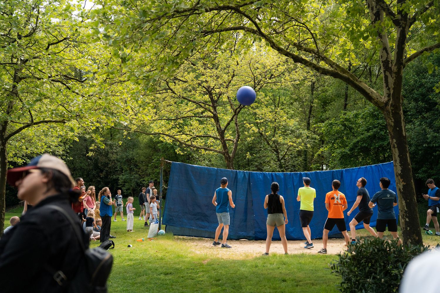 Group of young people play volleyball outdoors in a wooded park setting on a sunny day.