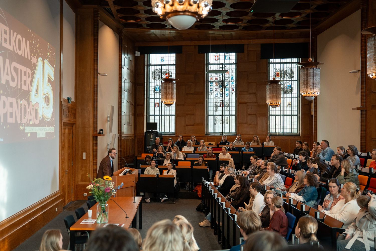 Crowded university master open day welcome presentation in historic wood-paneled lecture hall.