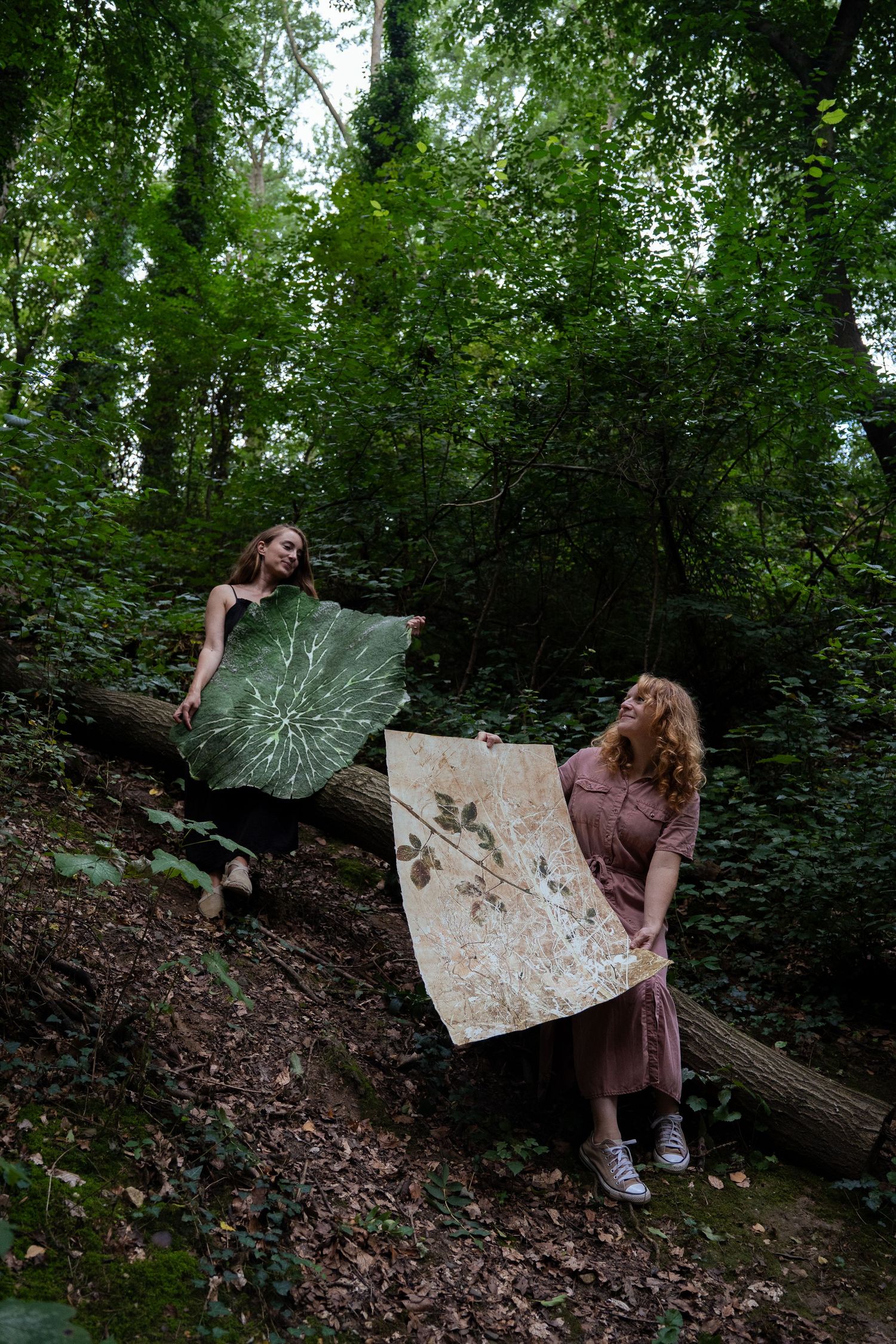 Two women displaying botanical print artworks in a lush green woodland forest.