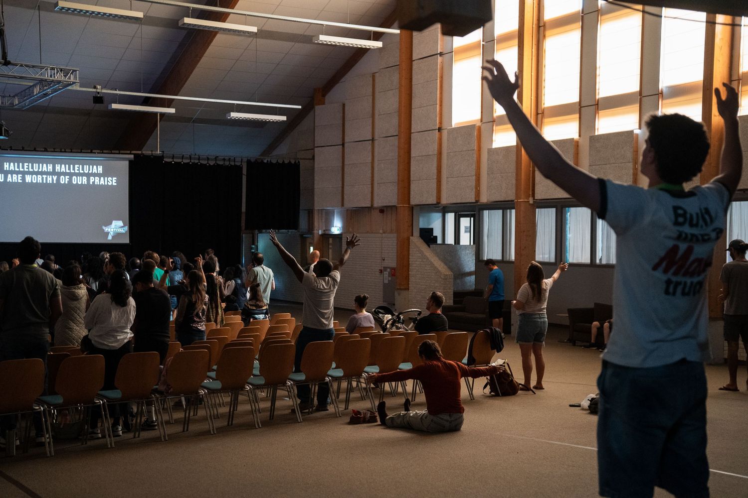 Young people raise hands in worship during a Christian praise gathering in a modern hall with projected lyrics.