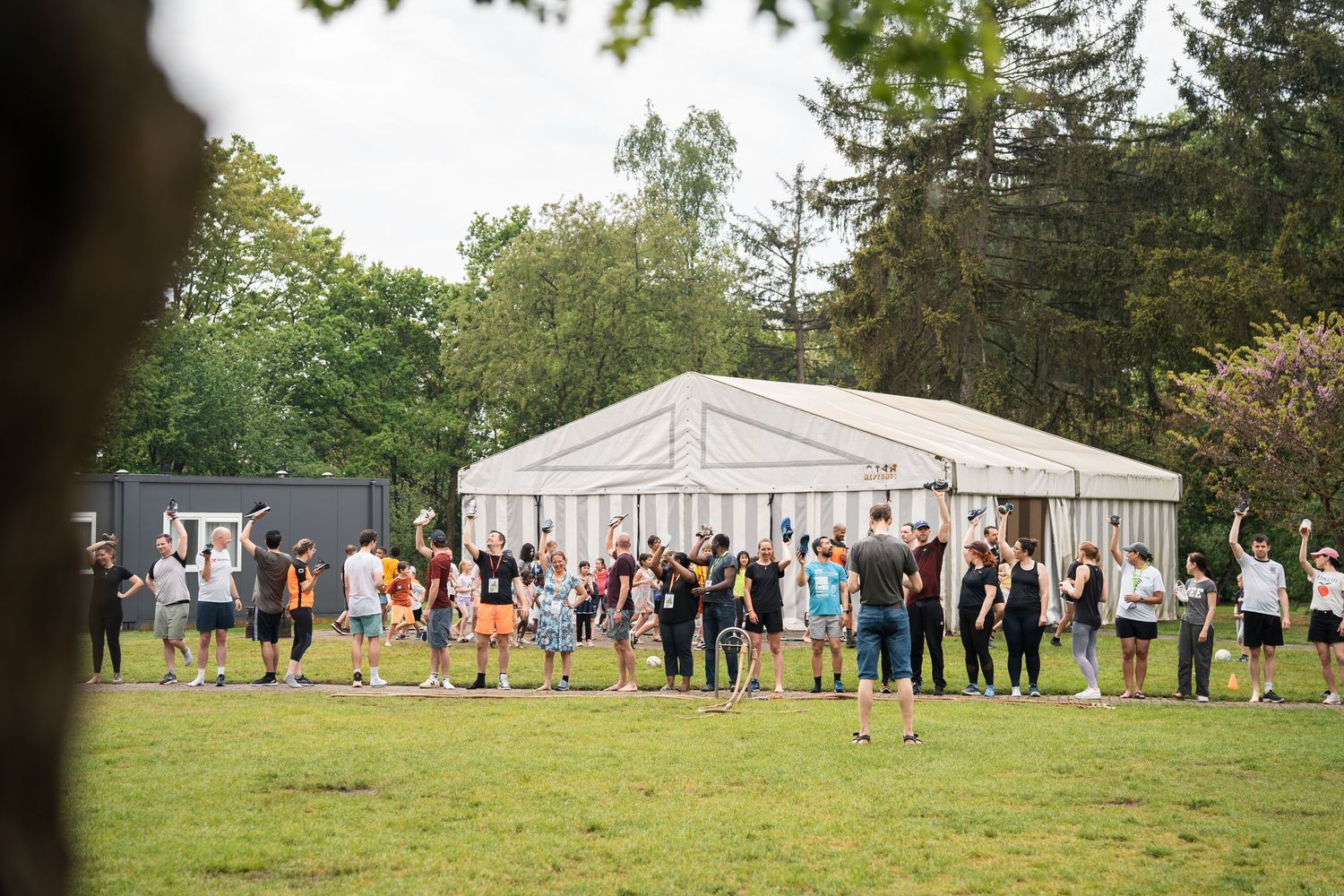 Large crowd gathered outside a white event marquee tent on green grass surrounded by tall trees.