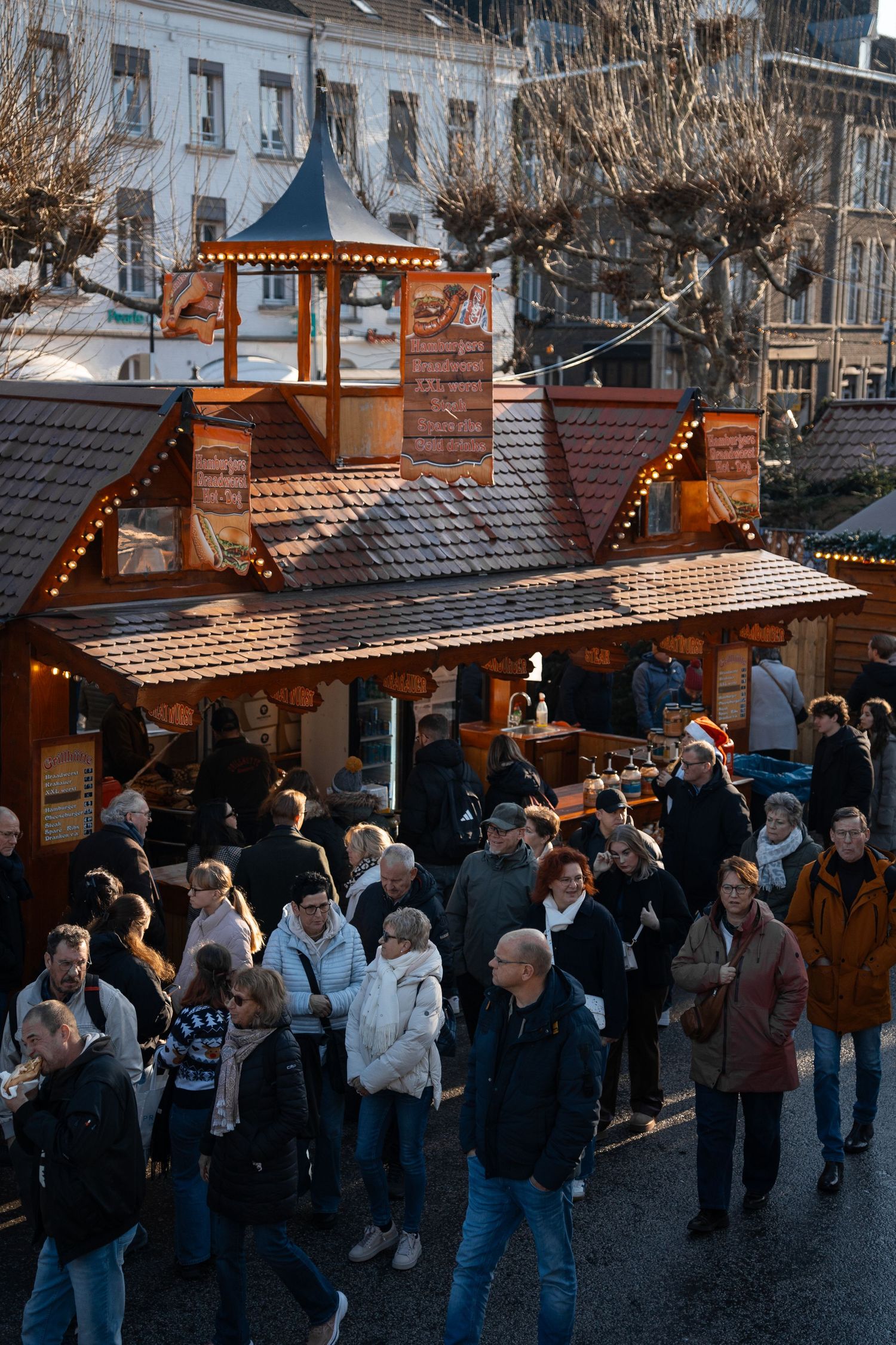 Aerial view of crowds browsing wooden stalls at a festive outdoor Christmas market with fairy lights.