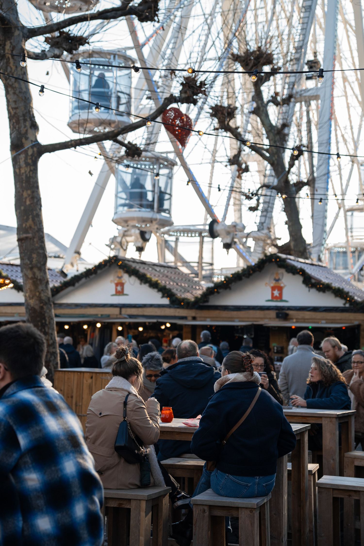 Visitors sitting at outdoor tables at a Christmas market with a large Ferris wheel in background.