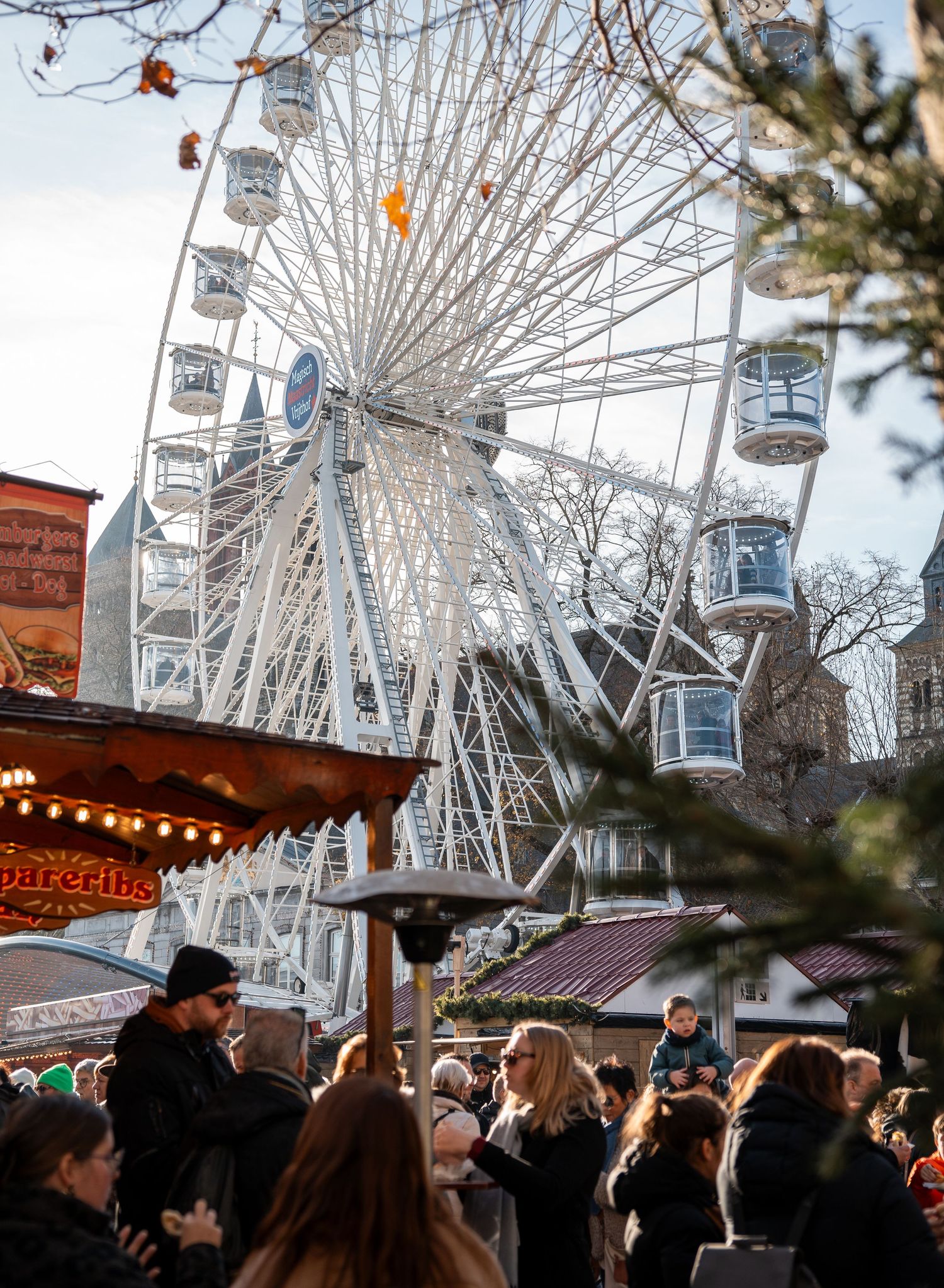 Busy Christmas market with a large white Ferris wheel towering above festive wooden stalls and crowds.