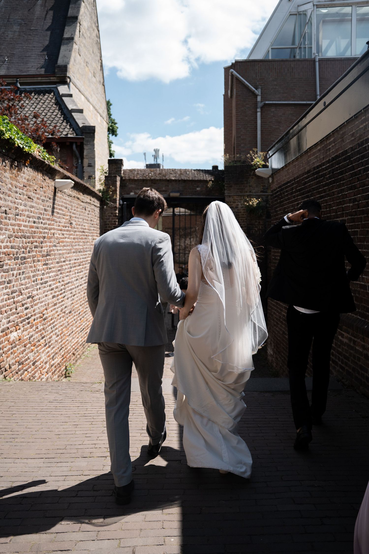 Bride and groom walk hand in hand through a brick alleyway in daylight, bride wearing a long veil.