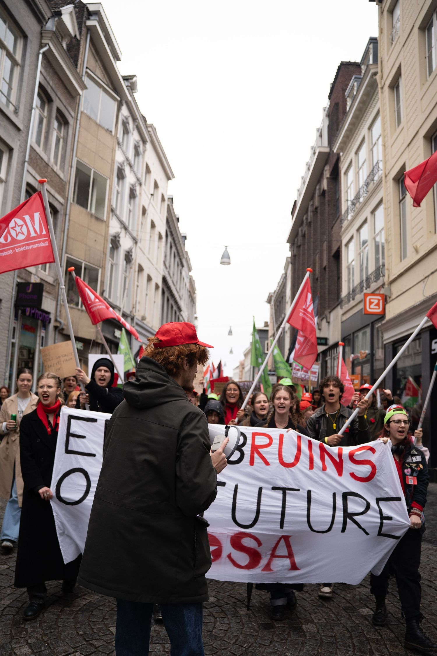Demonstration march through Maastricht city street with protesters holding Future Mosa banner and red flags.
