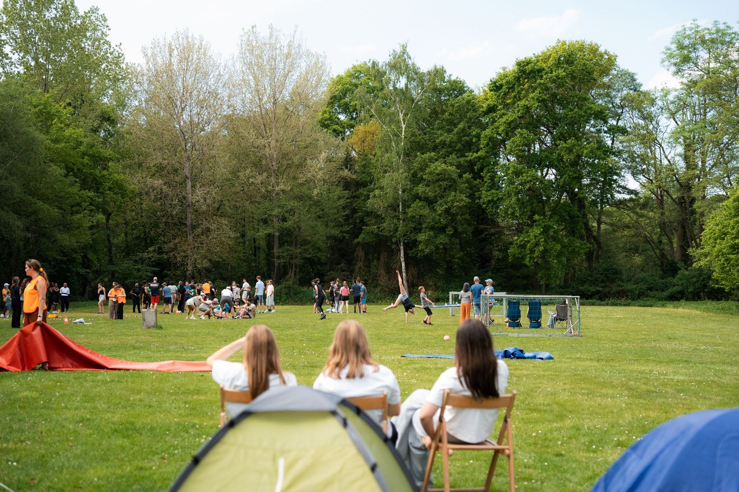 Children sit on folding chairs watching outdoor games on a green field surrounded by tall trees.