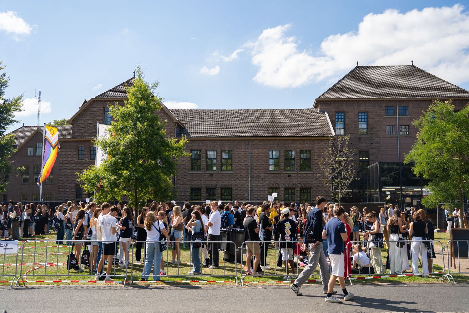 Large crowd of new students queuing outside brick university building on sunny day.