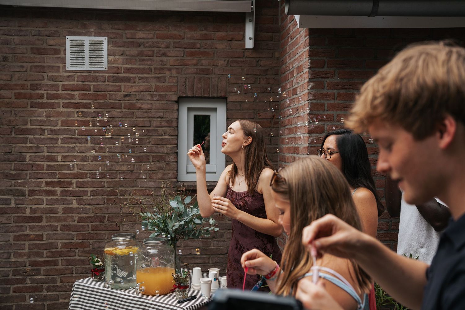 Young people socializing outdoors at a party near a brick wall, with a woman blowing bubbles near a drinks table.