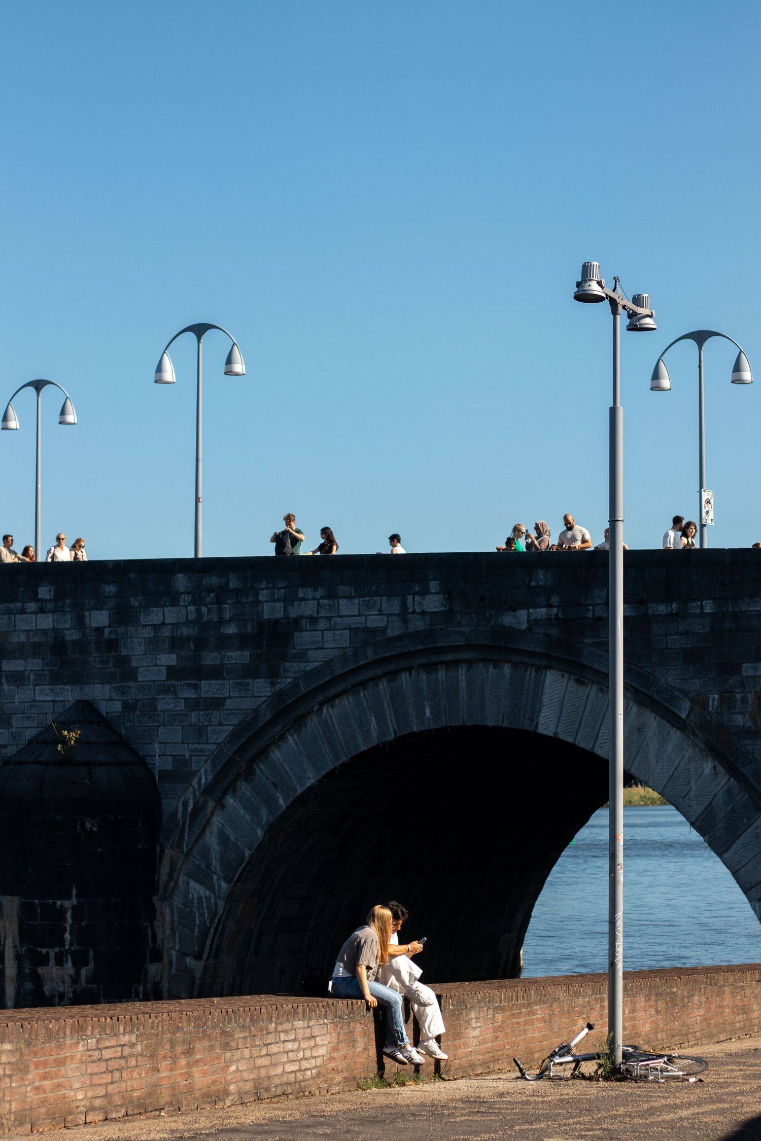 Couple sitting on a riverside wall beneath a historic stone arch bridge with people walking above.