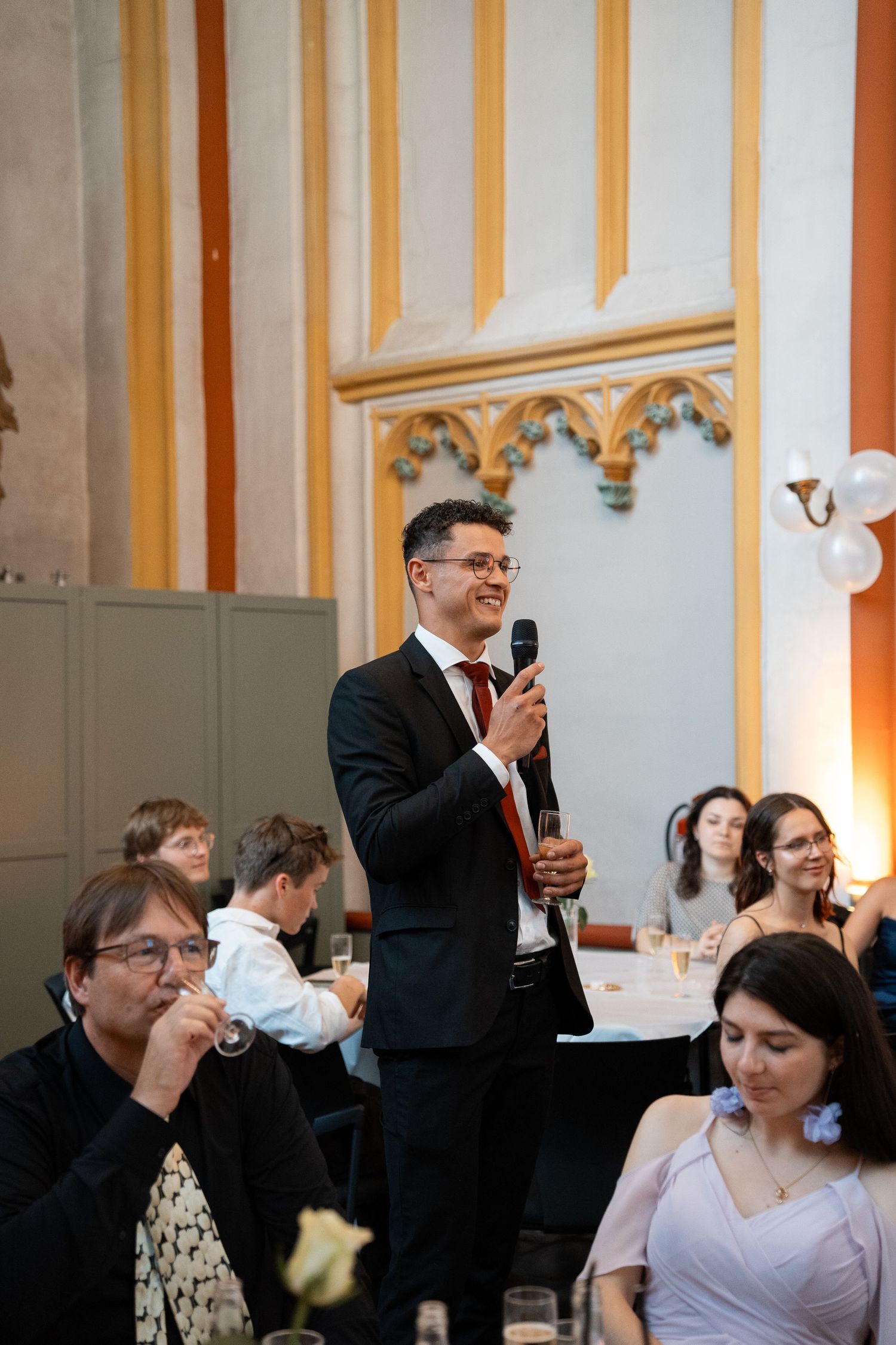A smiling man in a black suit gives a toast with a microphone at an elegant wedding reception.