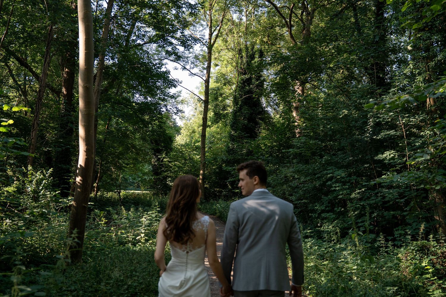 Bride and groom walking hand in hand through a lush green forest path.